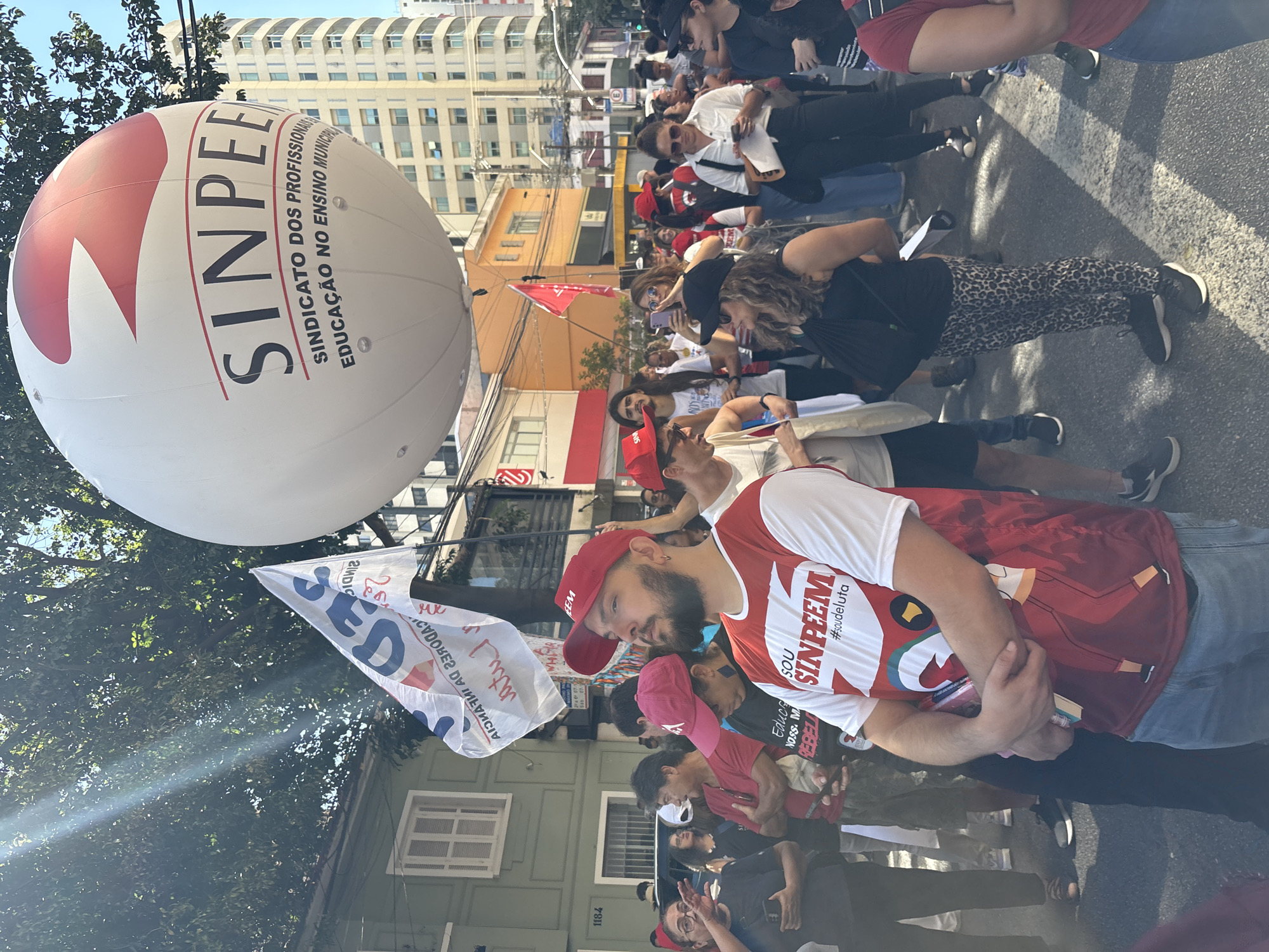 A street-level, eye-level shot depicts a demonstration or protest in a sunny urban environment, likely São Paulo, Brazil, based on the language and references in the signage.
A large, white balloon with the red logo "SINPEEM" (Sindicato dos Profissionais em Educação do Ensino Municipal) dominates the upper left of the frame. The phrase "SINDICATO DOS PROFISSIONAIS EDUCAÇÃO NO ENSINO MUNICIPAL" is written in a smaller font under the logo.
A crowd of people, presumably demonstrators, is visible. Most are wearing red clothing or carrying signs, suggesting solidarity with the SINPEEM union. One man in the foreground wears a red and white shirt with the "SOU SINPEEM #soudeluta" logo on it. He also sports a red cap.
Flags and banners are present, including one bearing the word "ZIMDIC".
The backdrop includes buildings of varying heights and styles, typical of an urban landscape. A building numbered "1184" is visible on the left.