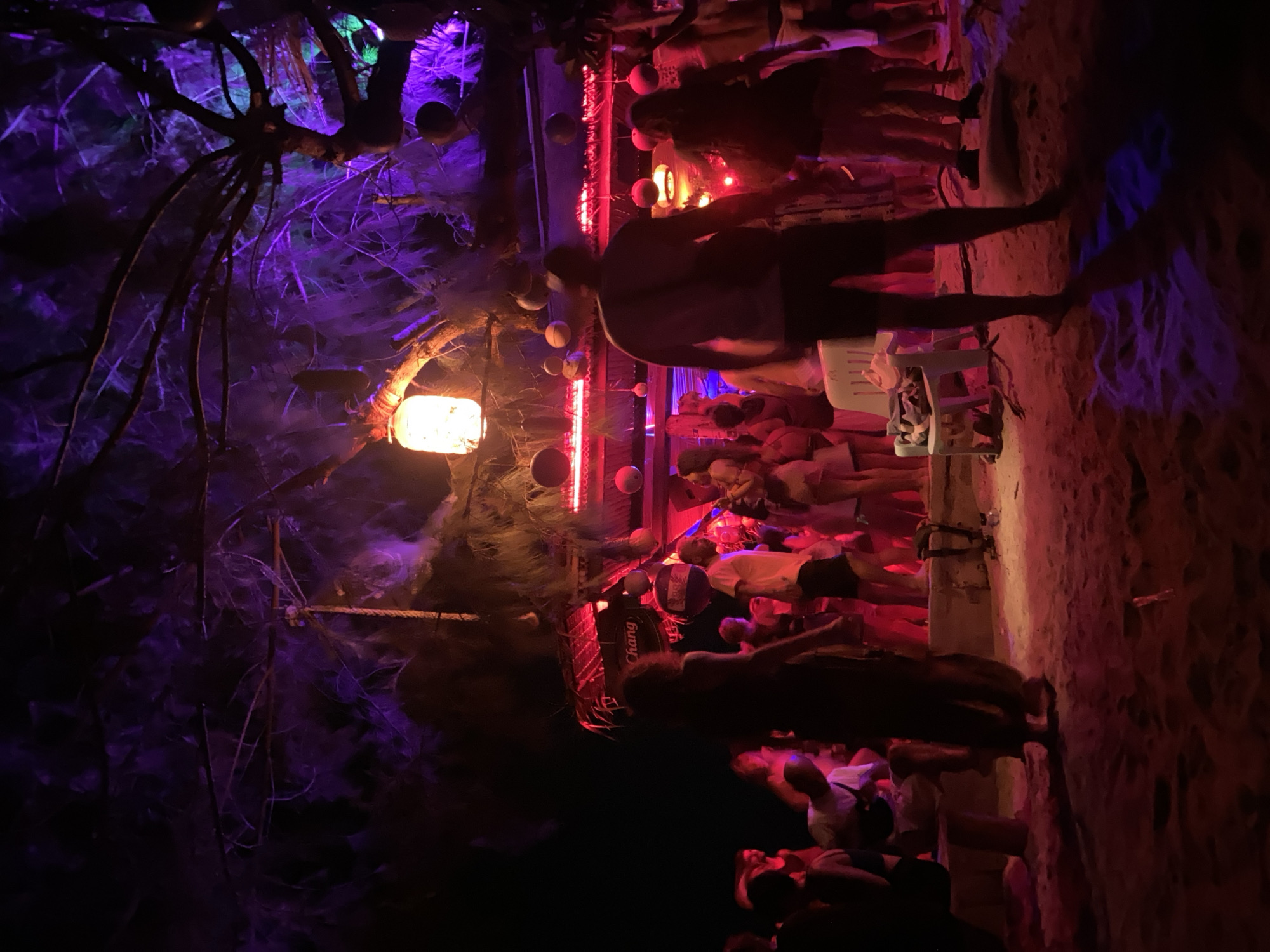 The image shows a nighttime scene at a beach bar in Baan Sri Thanu, Thailand. The bar area is illuminated with red and purple lighting, and a hanging lantern is visible amongst tree branches on the left. People are gathered, standing and sitting in the bar area. Some figures are silhouetted against the light. There is sand on the ground. A "Chang" beer sign is visible on the roof of the bar. Decorations such as balls are hanging in the bar area.