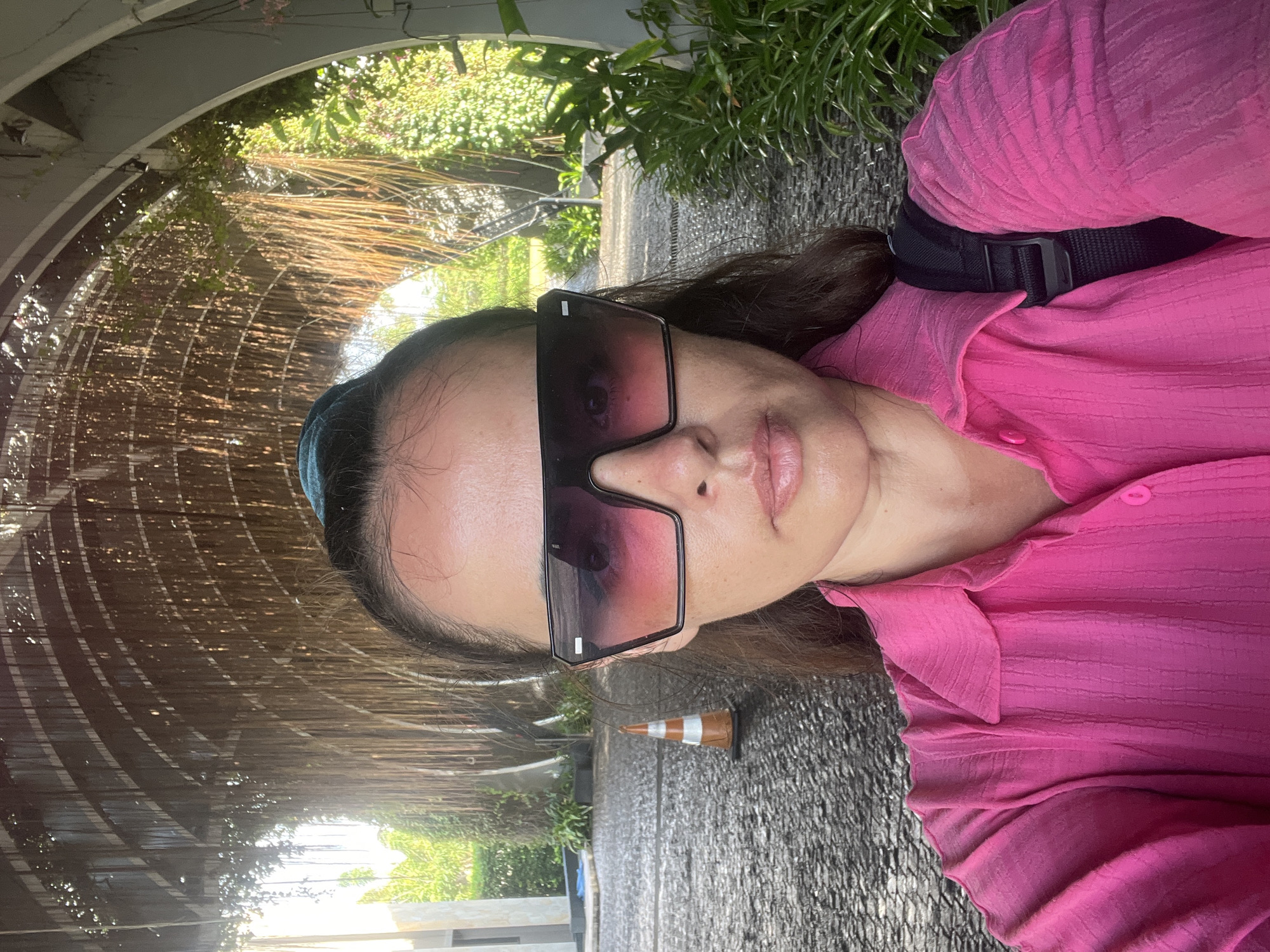 A woman is taking a selfie in front of an arched walkway covered in hanging vines. She is wearing large, angular sunglasses with a pink gradient lens and a bright pink collared shirt. Behind her, the walkway is paved with dark stones, and a traffic cone is visible on the left side. Lush green foliage lines the right side of the walkway, and sunlight filters through the vines overhead. The woman is positioned in the center of the frame, with her face and upper body taking up most of the image. The setting suggests an outdoor area, likely in a tropical or subtropical environment, possibly a resort or a public garden in Kuta Selatan, Indonesia.
