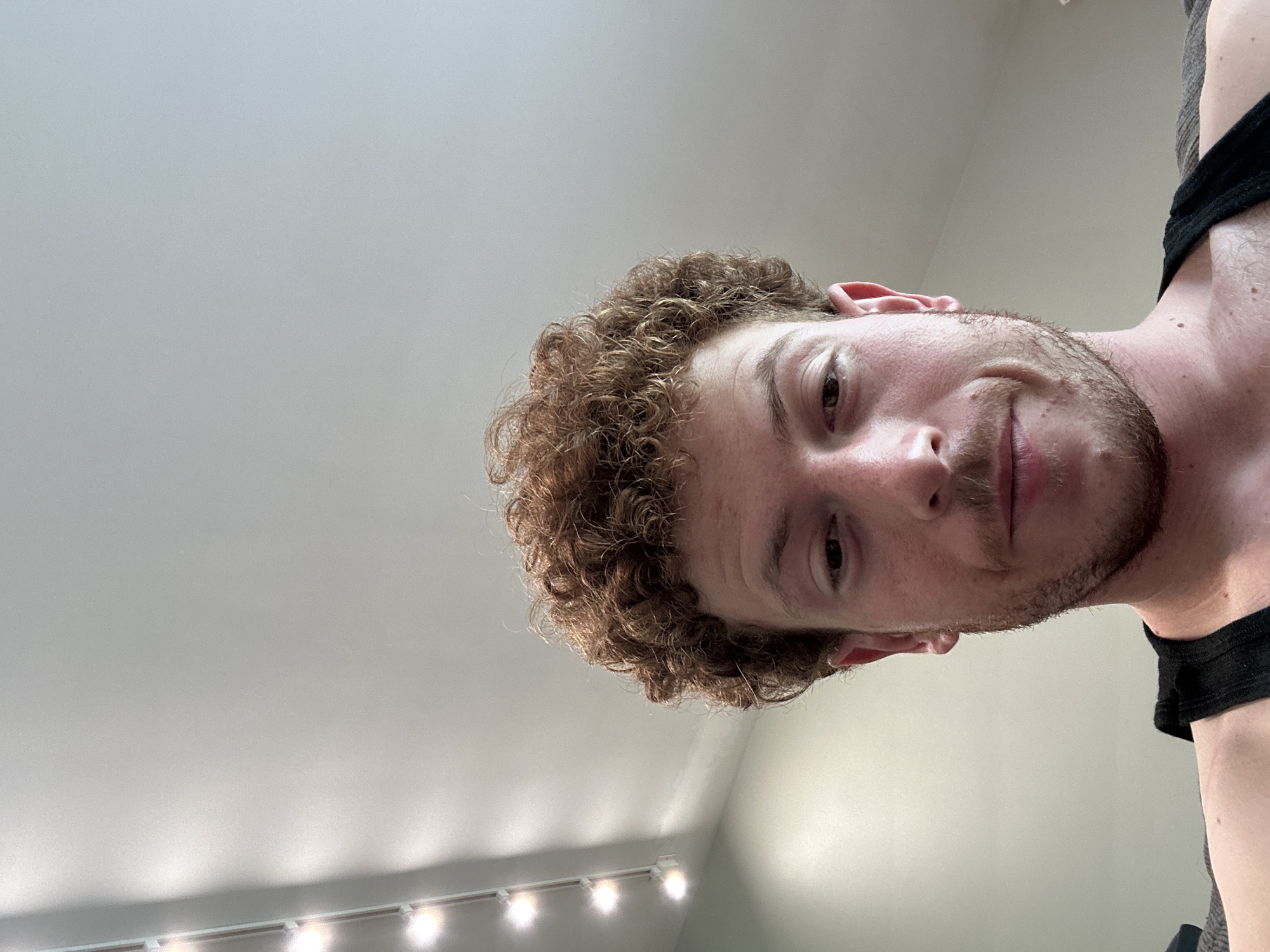 A person with curly, reddish-brown hair, stubble, and a dark tank top is pictured from a low angle. The background shows a ceiling with track lighting. The location is Pepper Pike, United States of America.