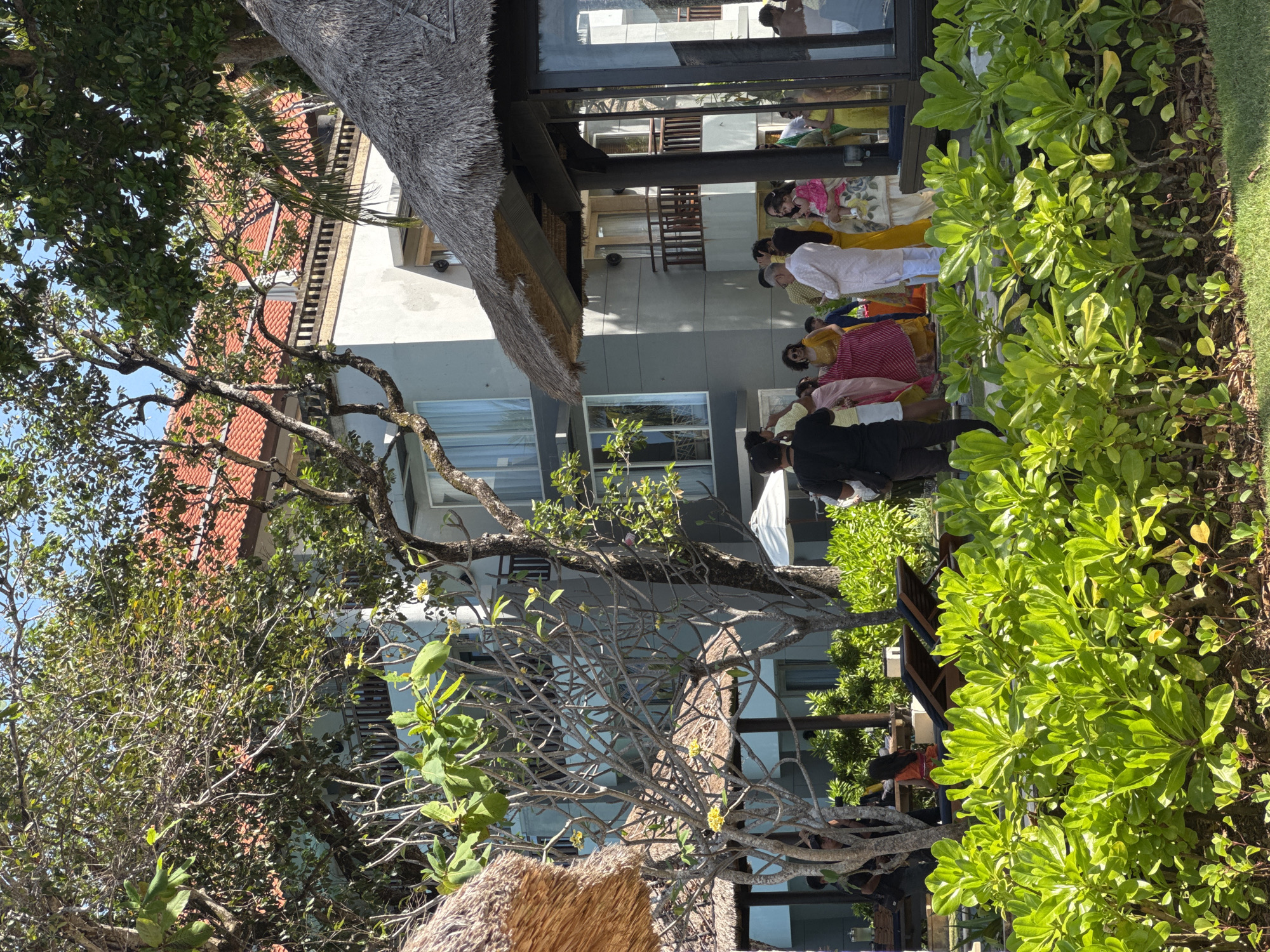 The image depicts a group of people, some wearing traditional clothing, gathered near a building in Kuta Selatan, Indonesia. The building features a thatched roof and modern architectural elements. Lush greenery and trees surround the area. The people appear to be engaged in some kind of event or ceremony.