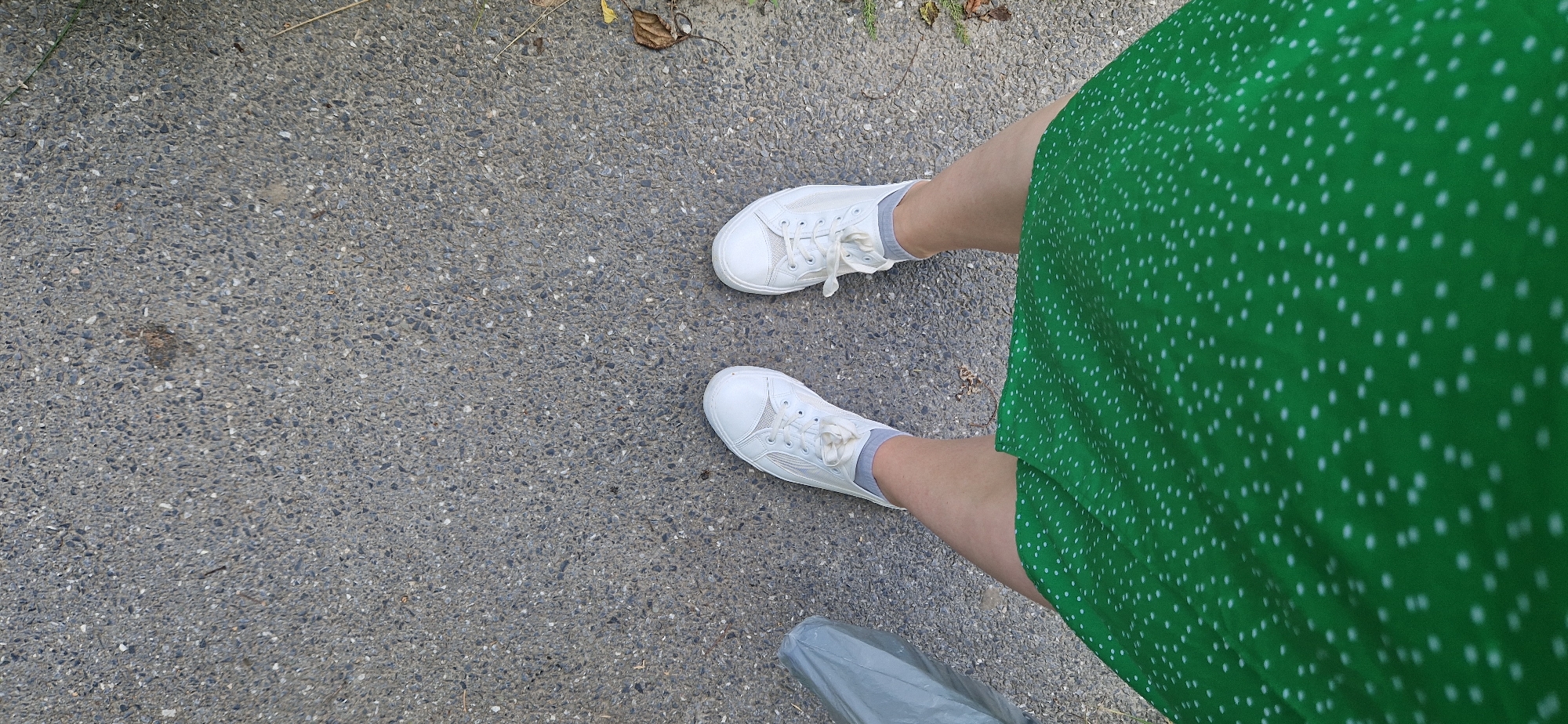 This is a top-down, full-frame shot of a person's legs and feet standing on a gray asphalt surface. The person is wearing white low-top sneakers with white laces and gray socks. They are also wearing a green knee-length dress with a white polka dot pattern. To the left of the person's feet is a gray plastic bag. The asphalt surface appears to be outdoors, possibly in a city, and shows some small pebbles and debris. There are also some dry leaves and grass visible on the right side of the frame.