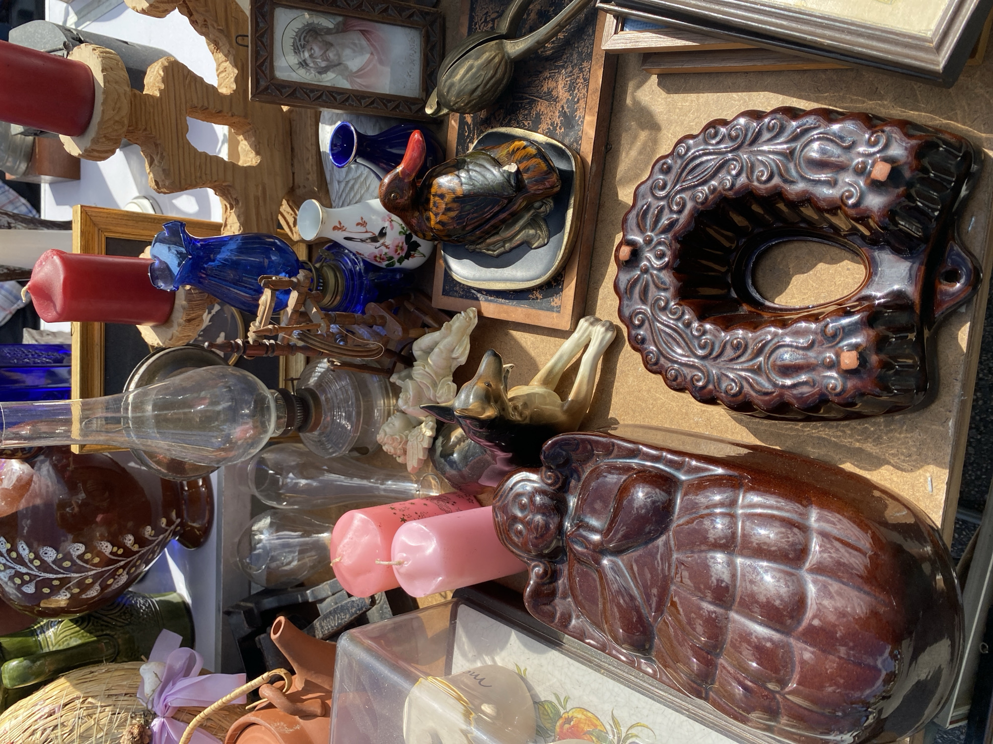 This image depicts a stall at an outdoor flea market, likely in Vienna, Austria, given the style of the ceramics. The stall is laden with various antique and decorative items. Prominently featured are several ceramic pieces: a large, dark brown, glazed ceramic mold shaped like an ornate ring cake or bundt cake, and a long, similarly glazed brown ceramic piece resembling a reclining figure with hands clasped.
Other objects on display include a glass oil lamp with a metal base, two pink pillar candles, and two red pillar candles on wooden candle holders. There's a small blue glass vase, a white ceramic vase with floral decorations, and a dark blue ceramic vase. A small, framed picture of Jesus with a crown of thorns is visible, alongside a decorative ceramic duck figurine with a red beak, resting on a dark plate.
Further back, a large, patterned brown vase, a woven basket, and a small terracotta teapot are visible. Several clear glass items, possibly vases or bottles, are also present. A dog figurine, appearing to be made of metal or ceramic, is positioned near the center of the display. The scene is set outdoors, with a table or surface covered in a brown cloth, and sunlight casting shadows. The overall impression is one of a curated collection of vintage and decorative items for sale.