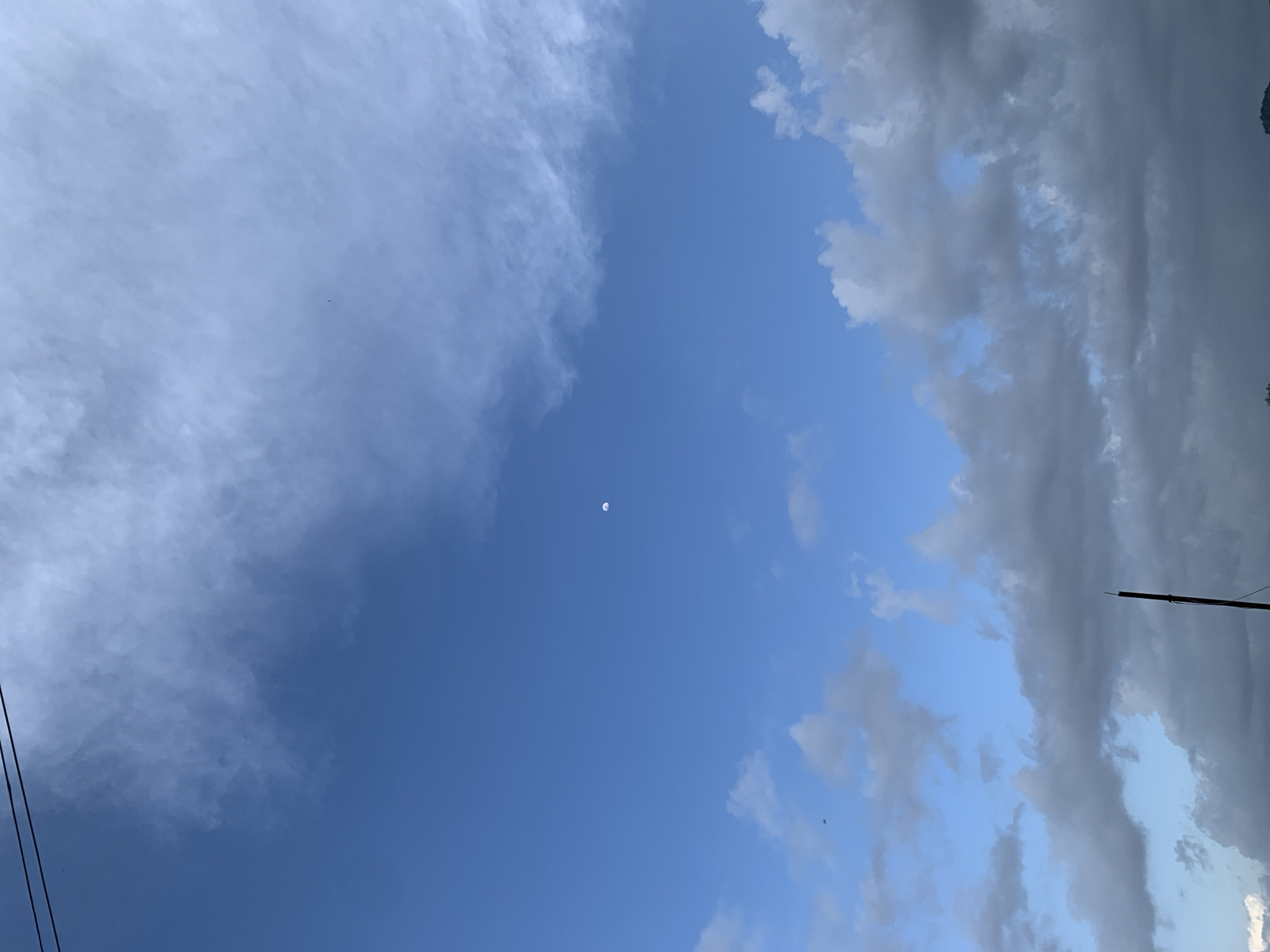 The image shows a bright blue sky with scattered clouds. The moon is visible as a small crescent in the upper right portion of the sky. There are also two thin, dark lines stretching across the top of the image, which appear to be power lines. In the lower portion of the image, there are some dark clouds. A dark, vertical object, possibly a pole or antenna, is visible in the lower right corner. The image depicts an outdoor scene, likely taken in Kuru Karama, Nigeria, given the context provided.