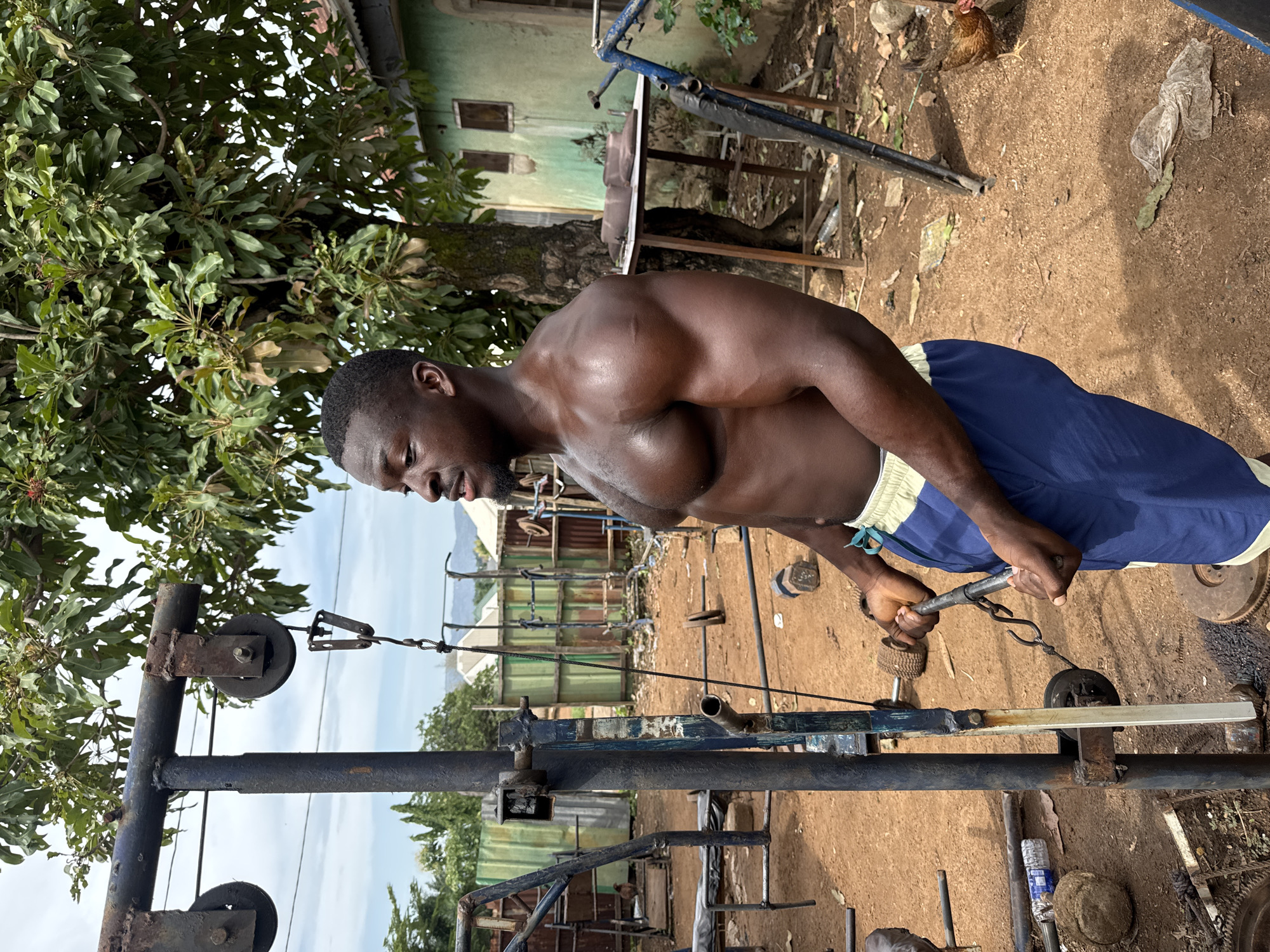 A man is working out outdoors in Akwanga, Nigeria. The location appears to be a makeshift gym area with various pieces of equipment. He is shirtless, wearing blue shorts with a light-colored waistband, and is pulling on a weight machine cable. The environment includes trees, a building in the background, and loose dirt. A chicken is present in the background. Other pieces of exercise equipment are scattered around the scene.