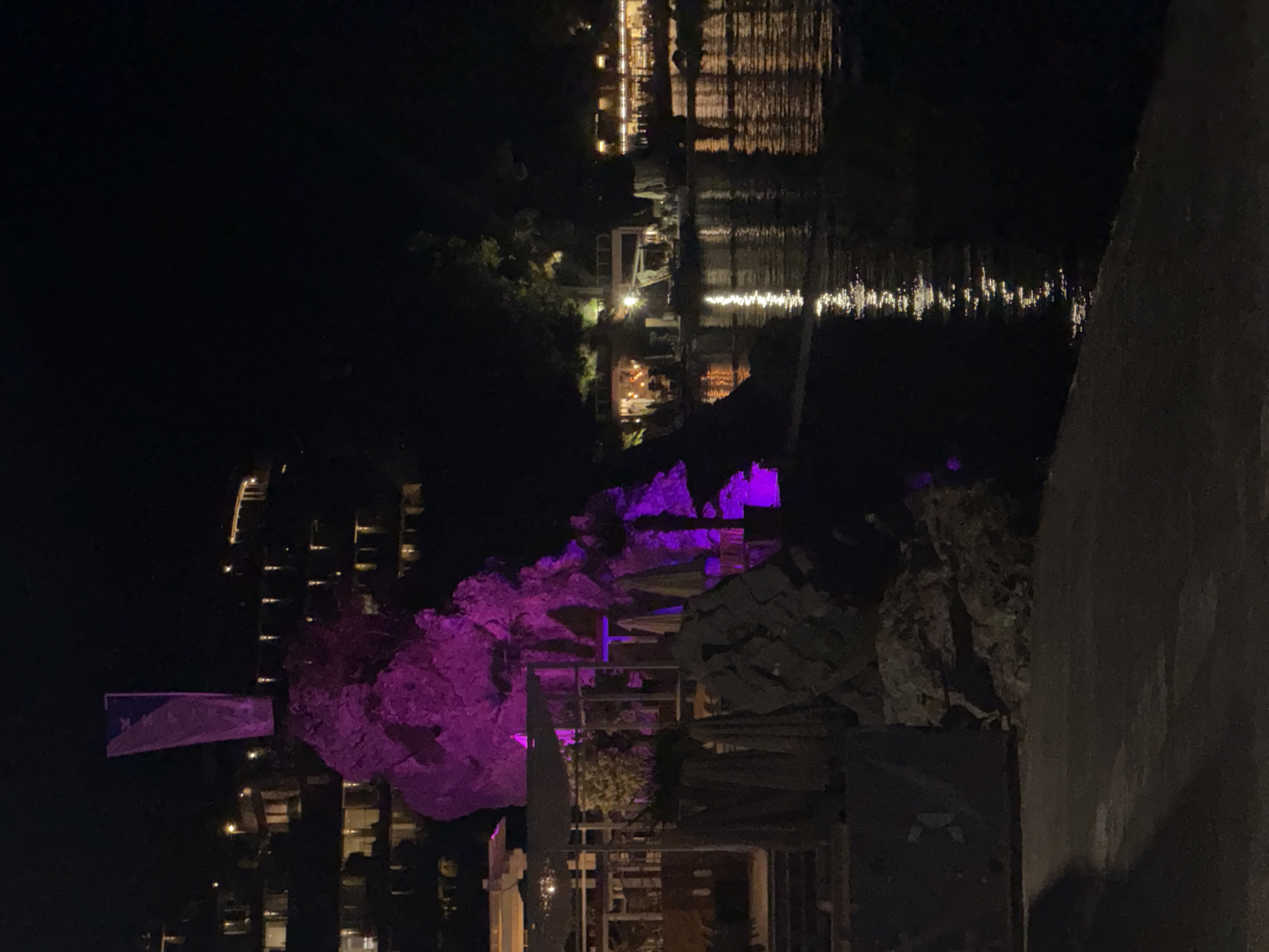 The image captures a nighttime scene in Budva, Montenegro, along the waterfront. In the foreground, a dark, paved walkway runs along the edge of a body of water, likely the Adriatic Sea. To the left, a rocky outcrop is dramatically illuminated with vibrant purple lighting, highlighting its textured surface. A flag or banner, also illuminated with a soft purple hue, is affixed to the top of the rock formation.
Behind the illuminated rocks, a multi-story building or complex is visible on a hillside. Its windows are lit, suggesting occupancy. Further to the right, across the water, a strip of lights indicates a row of buildings or establishments, possibly restaurants or hotels, bordering the water's edge. The lights from these buildings are reflected on the dark surface of the water, creating shimmering streaks of light. The overall atmosphere is one of a lively, illuminated nighttime resort or coastal town.