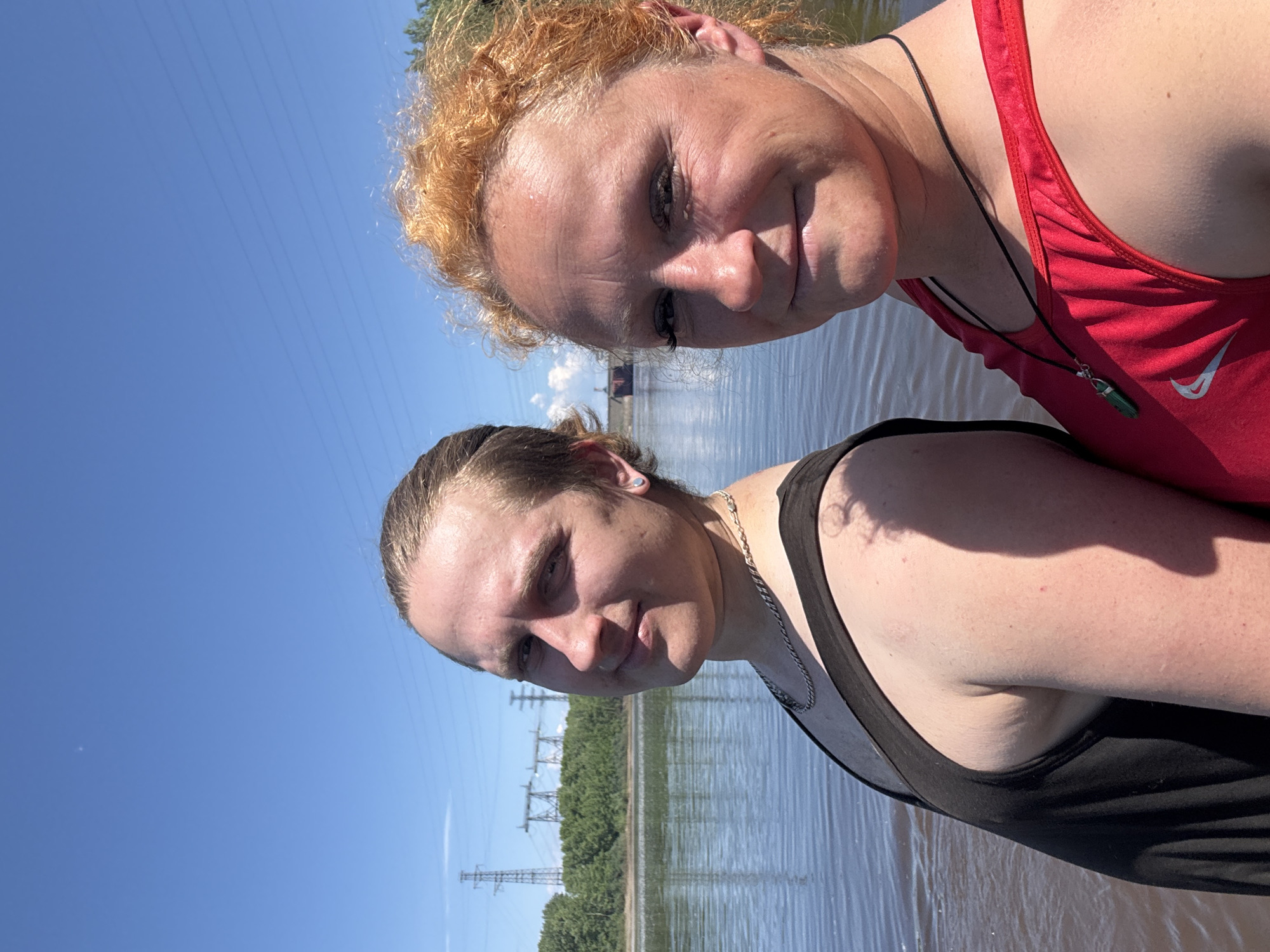 Two people, one with blonde hair tied back and wearing a black tank top and a silver chain, and another with reddish-orange curly hair and wearing a red tank top with a green pendant, pose for a selfie in front of a body of water. The person on the left is looking towards the camera with a slight smile, while the person on the right is smiling. In the background, trees line the shore, and power lines and pylons are visible against a clear blue sky. The location is the Kama River in Chaikovsky, Russia.