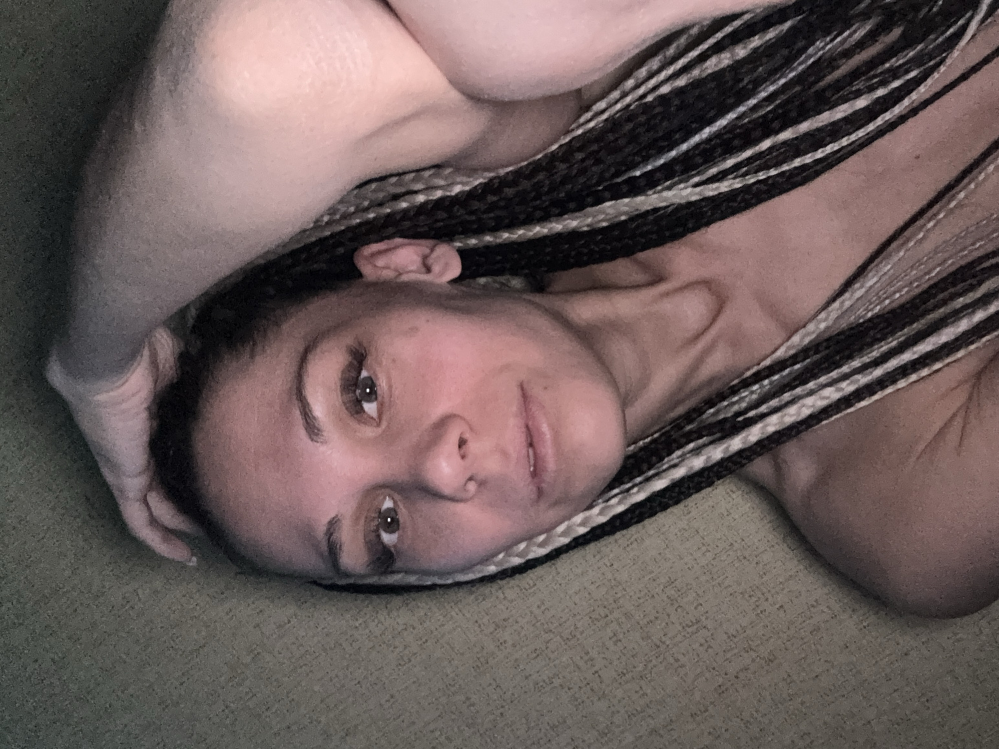 An intimate, high-angle portrait features a person, likely a woman, lying down on a soft, textured surface. Her head and upper torso are visible, positioned diagonally across the frame. She gazes directly towards the viewer with a neutral to slightly contemplative expression. Her eyes are brown, and her lips are slightly parted.
Her dark hair is styled in numerous long box braids, intricately woven with contrasting white or light-colored synthetic extensions. These braids cascade around her head and shoulders. One arm, possibly her left, is bent and resting with her hand near her head, partially visible against the textured background. Her other shoulder and upper arm are also visible, suggesting she is lying on her side or back.
The background is a consistent, light green or olive-green textured fabric, resembling upholstery or a blanket, which creates a soft, diffused setting. The lighting is gentle and even, highlighting her facial features without harsh shadows, indicative of an indoor environment. There are no other discernible objects, people, or text in the image. The overall impression is one of calm and casual intimacy. The provided context suggests the location is Rusanovo, Russia, though no specific visual cues confirm this.