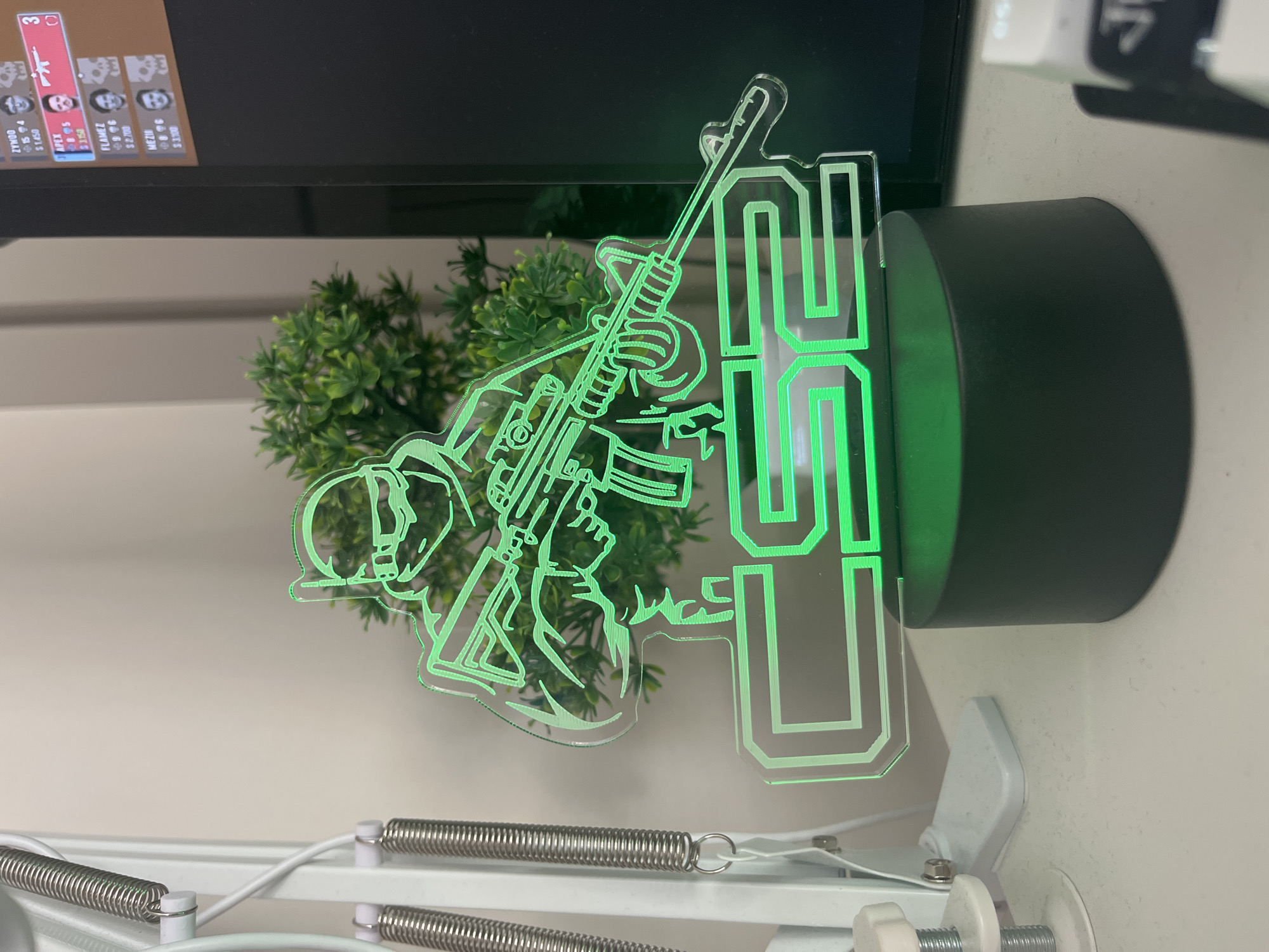 An acrylic lamp featuring a silhouette of a soldier holding a rifle and the text "CS2" glows green. The lamp is positioned on a white desk next to a potted plant and a monitor displaying a video game interface. A portion of a desk lamp with coiled springs is visible on the left. The scene is indoors, with the monitor suggesting a gaming setup. The image does not provide any information about the city of Varna, Bulgaria, or any specific location within it.