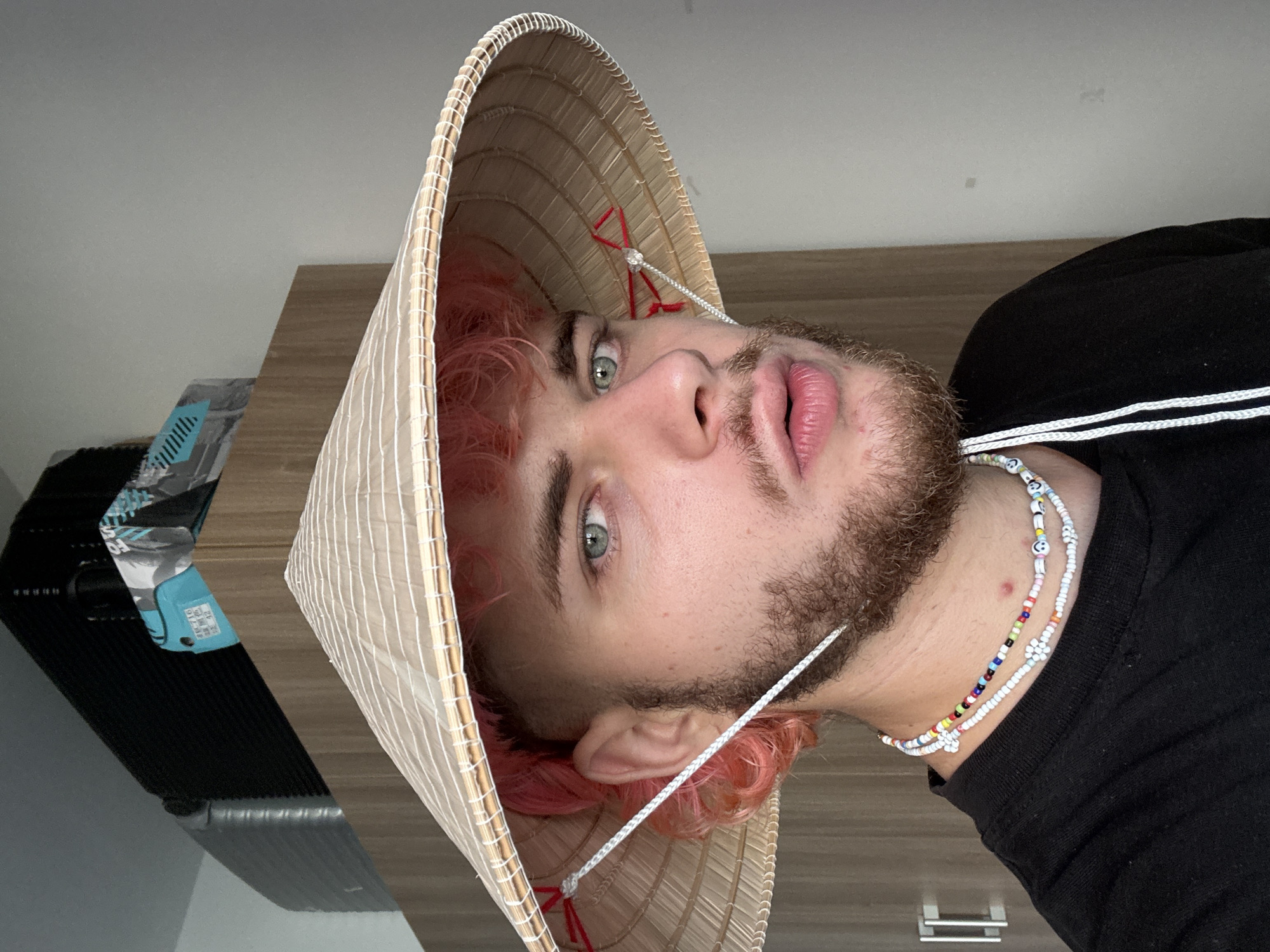 A medium close-up shot shows a person wearing a traditional Vietnamese conical hat, known as a "nón lá". The person has light skin, pink hair, and a beard. They are wearing a black shirt and two beaded necklaces. The background includes a wooden surface and a cooler. The location cannot be determined as being Đà Nẵng, Vietnam, from the visible elements.