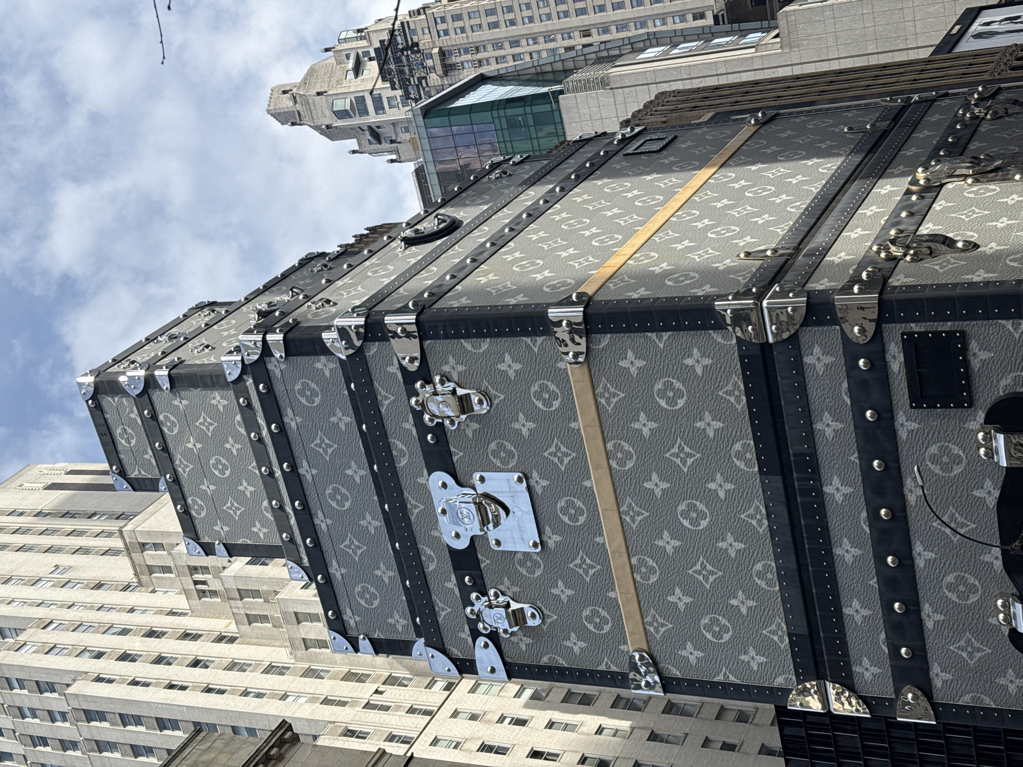 The image shows a large Louis Vuitton trunk placed on display outdoors in New York City, USA, with surrounding buildings visible. The trunk is rectangular and covered in the classic LV monogram pattern in shades of gray. It has metal reinforcing strips along its edges and corners, secured with rivets. The trunk features prominent metal latches on one side and a light-colored strap running vertically along its surface. The buildings surrounding the trunk are typical of urban architecture, featuring numerous windows and varying heights. The sky is partly cloudy.