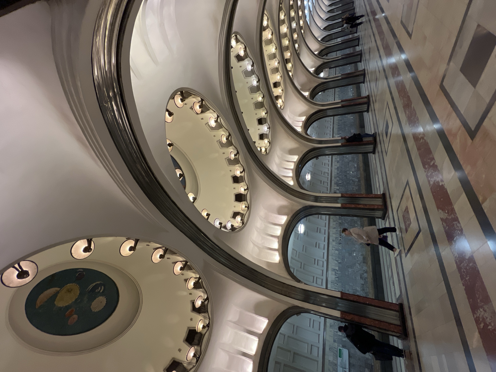 The image captures the interior of an underground metro station. The station is characterized by its repeating, arched structures with oval openings along the ceiling, each equipped with multiple light fixtures. A decorative circular artwork is visible on one of the ceiling arches.
Several people are present in the scene, appearing as they move along the platform. The floor is tiled with a pattern of rectangles, creating a striped effect. The arches appear to divide the platform, with a light-colored wall visible through each archway.