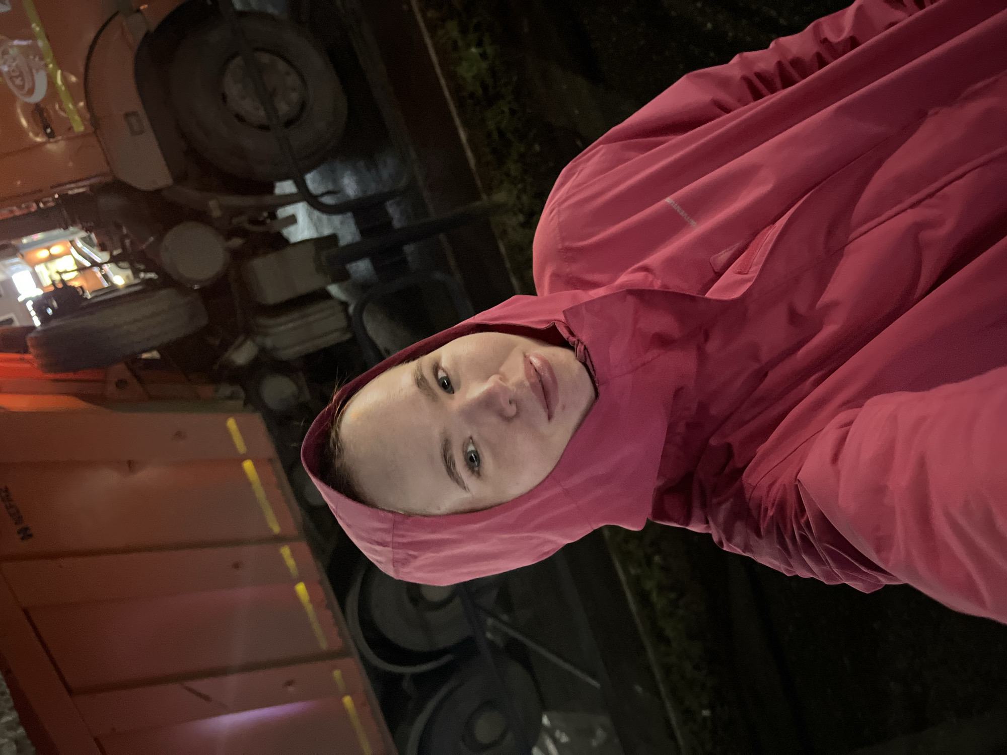 A person wearing a pink hooded jacket is standing next to a large orange truck, possibly a dump truck. The scene appears to be at night, as indicated by the darkness and artificial lighting. The location is identified as Chaikovsky, Russia. Details beyond these, like the exact reason for being near the truck, are not possible without additional context.