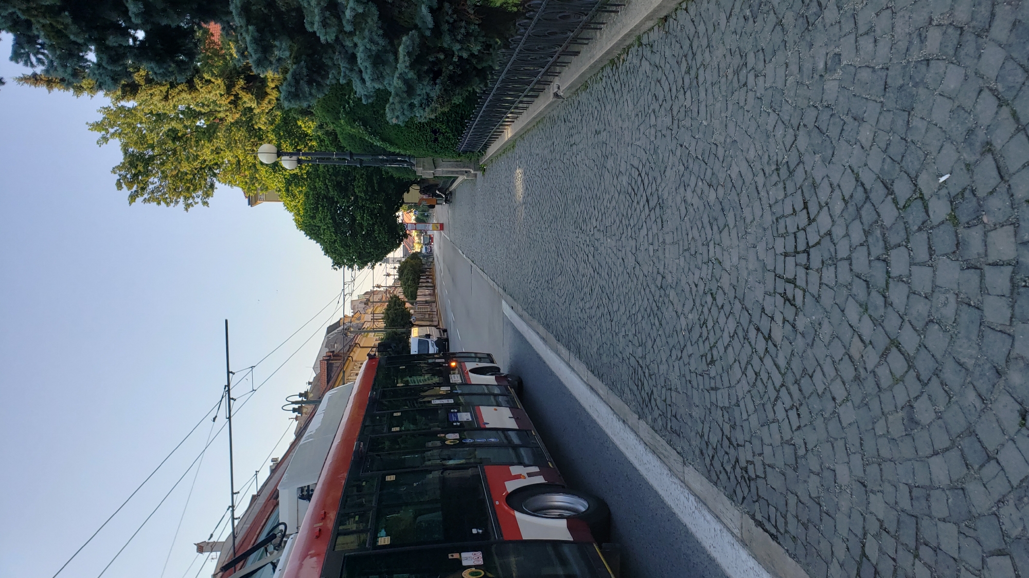 The image depicts a street scene in Prešov, Slovakia, with a red and white trolleybus on the left side of the frame, traveling on a road. The road is bordered by a cobblestone sidewalk on the right. Overhead wires for the trolleybus are visible against a clear sky. The sidewalk is lined with trees, a black metal fence, and lampposts. In the background, buildings with reddish roofs and more trees can be seen, suggesting an urban environment. The light suggests it is either early morning or late afternoon. No people are clearly visible or identifiable in the scene.