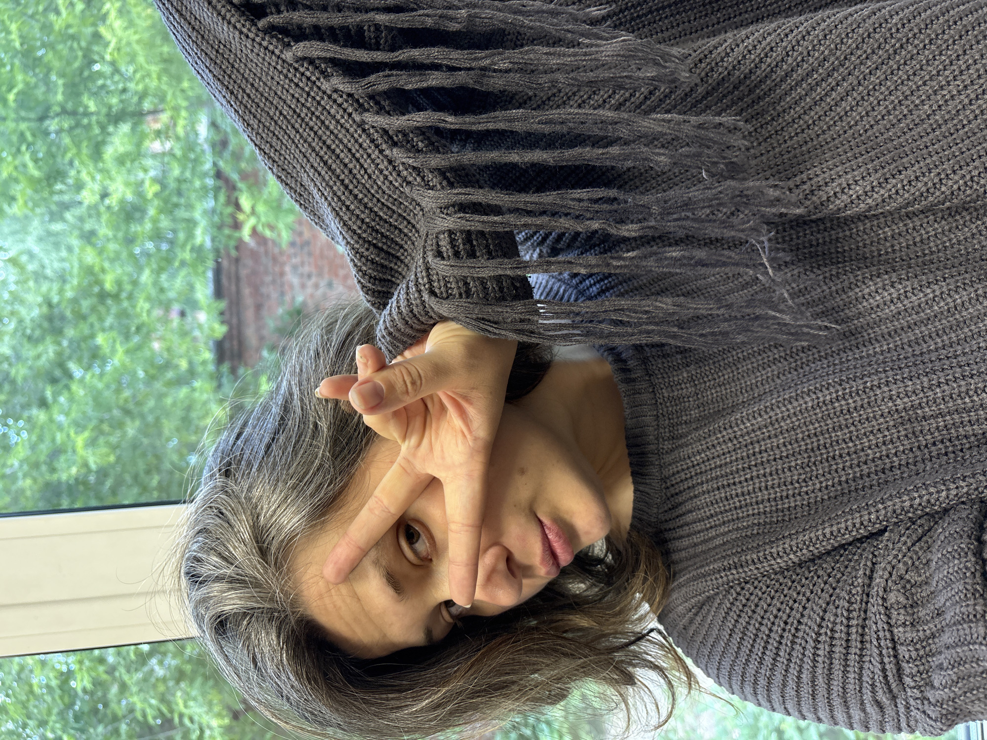 A young woman with grey-streaked brown hair is taking a selfie by a window. She is wearing a dark grey knitted sweater with fringe on the sleeve, and her right hand is raised to her face, forming a peace sign over her right eye. Her left arm is extended, also showing the fringed sleeve of the sweater. The background is out of focus, showing green foliage and a brick building, suggesting an outdoor setting or a view from a building. The image appears to be taken indoors, possibly in an apartment or office, given the presence of a window and interior lighting. There is no information in the image to indicate its location in Chaikovsky, Russia.