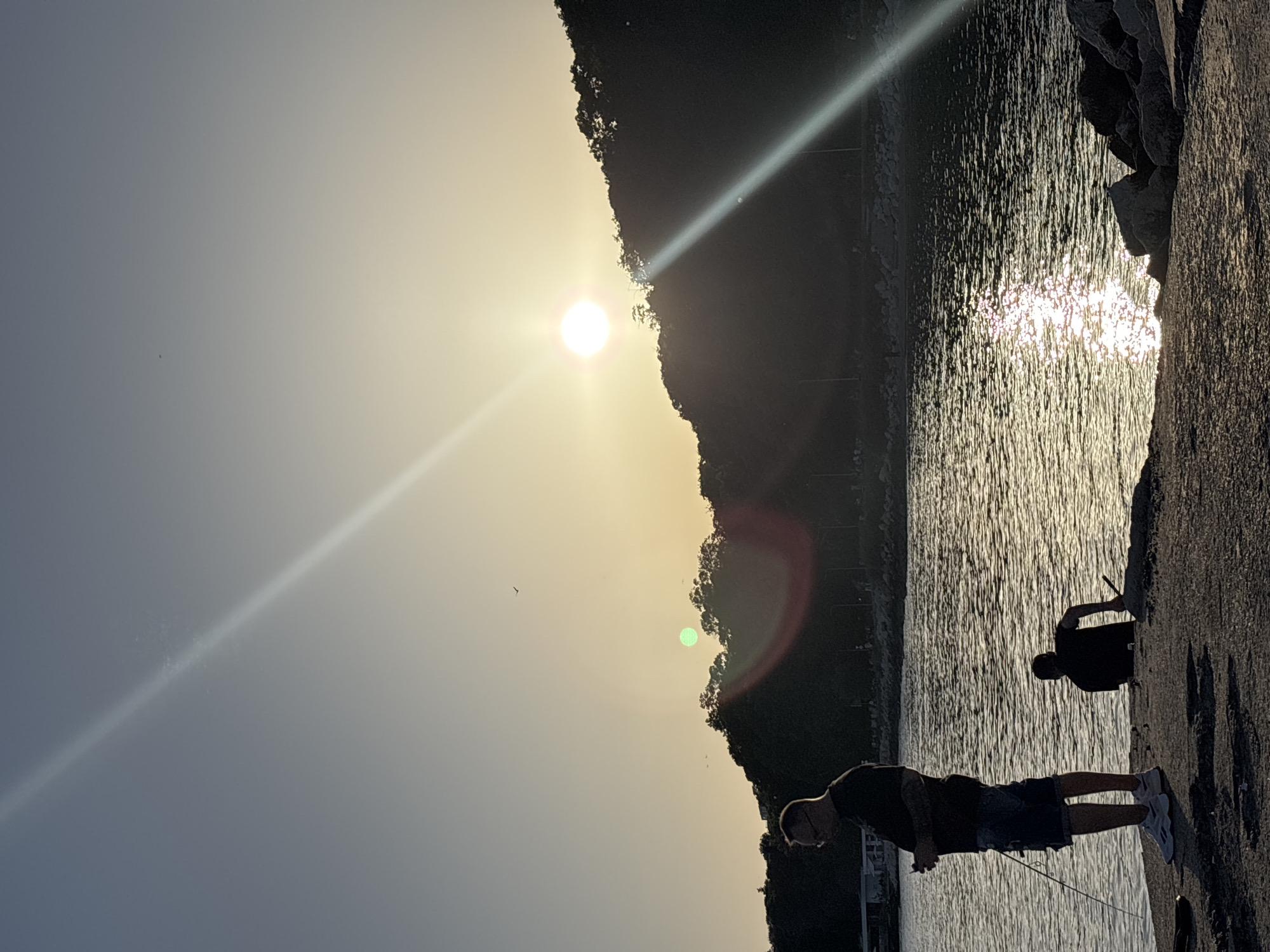 The image depicts a serene outdoor scene in Varna, Bulgaria, during what appears to be a sunset or sunrise. The sun is low in the sky, casting a warm, golden glow and creating strong lens flare effects that streak across the frame. The scene is set by the water, likely a pier or a rocky shoreline, with a dark, silhouetted hillside covered in trees in the background.
Two individuals are visible. In the foreground, a person wearing a t-shirt and shorts stands facing the water, holding what appears to be a fishing rod. Further back, another person, also in silhouette, is seated or leaning, also seemingly engaged in fishing. The water is calm, with shimmering reflections of the sun's light creating a textured surface. The rocky embankment on the right provides a natural foreground element. The overall atmosphere is peaceful and tranquil, suggesting a quiet recreational activity by the water. The time of day is clearly indicated by the low angle of the sun and the quality of the light. No visible text is present in the image.