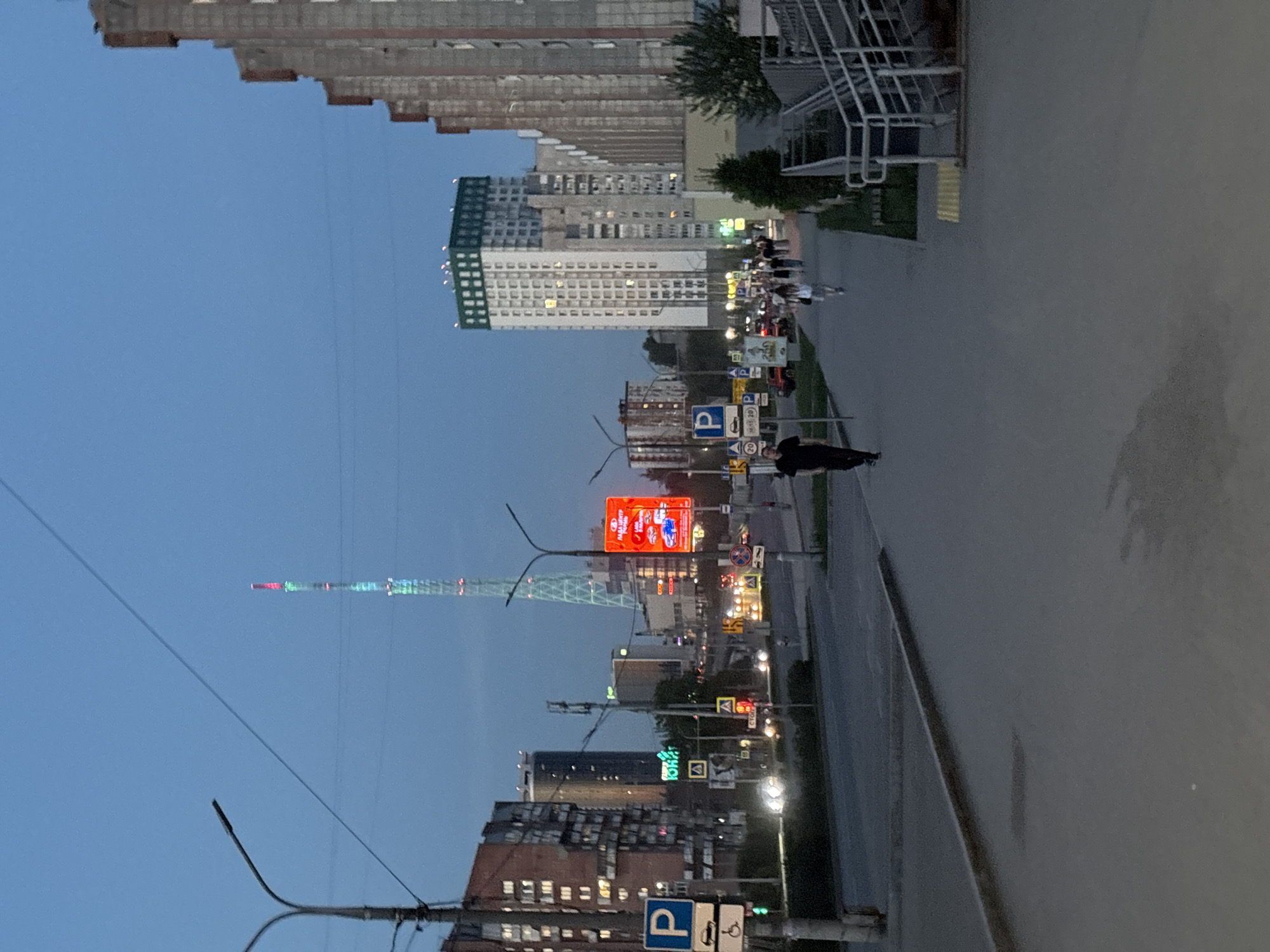 The image shows a street scene in Perm, Russia at dusk. Tall apartment buildings are visible in the background, with the Perm TV Tower featuring red and green lights. A large red electronic billboard is located further down the street. Streetlights illuminate the road, where a person is walking on the sidewalk. Various street signs are visible along the road.