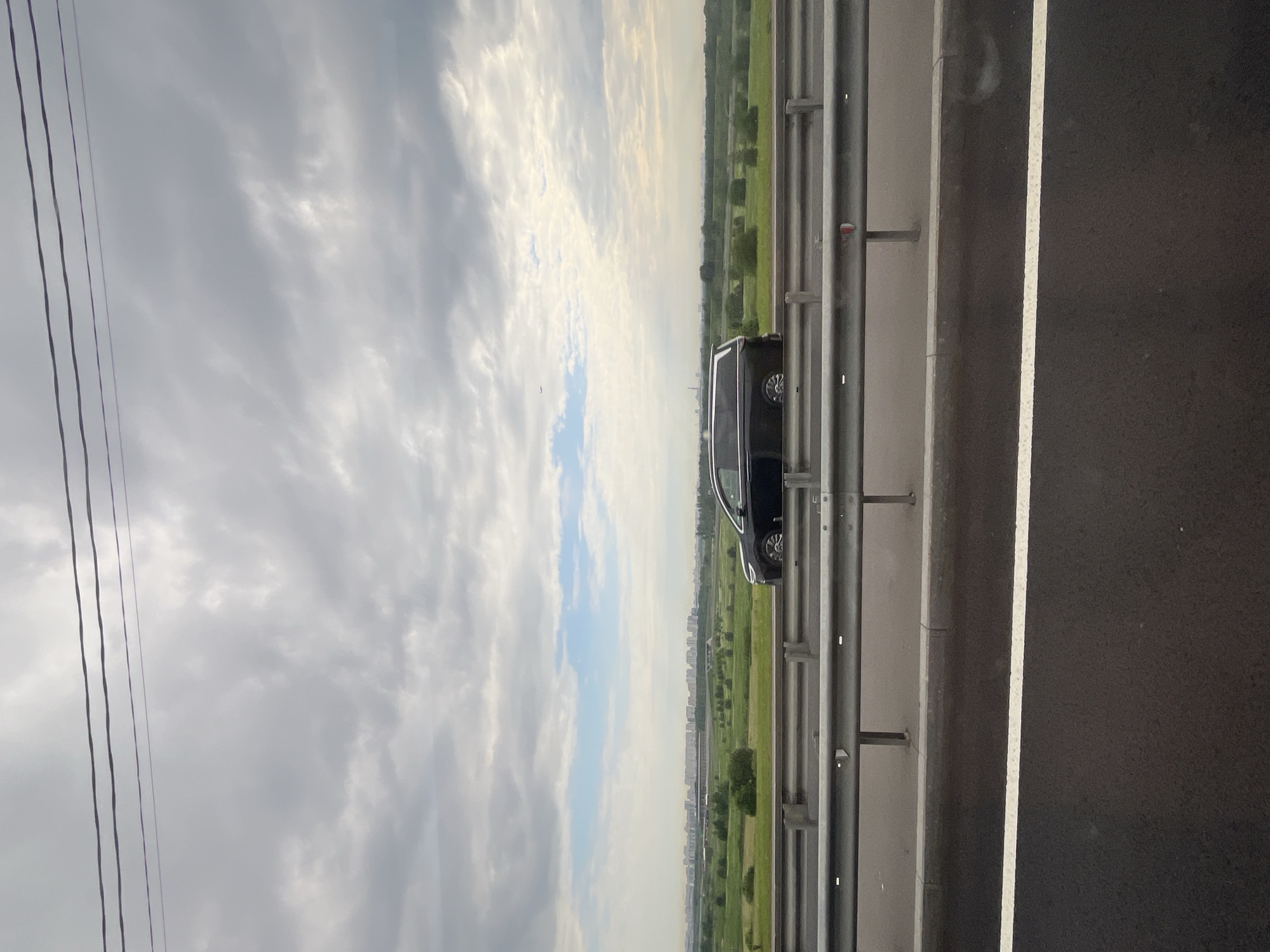 The image presents a wide, horizontal landscape view, likely captured from a moving vehicle on a highway. The upper half of the frame is dominated by a vast, partly cloudy sky, featuring a mix of grey and white clouds with visible patches of brighter blue. In the top-left corner, three dark power lines stretch diagonally across the sky.
Below the sky, the mid-ground reveals an expansive green landscape with fields and scattered trees, extending towards a distant horizon. A faint, blurred outline of buildings, suggesting a city or urban area, is visible on the horizon. A tiny dark speck, possibly a bird or a distant aircraft, is discernible in the sky above the distant cityscape.
The lower right portion of the image shows a dark asphalt road surface, marked by a clear white solid line that delineates a lane or shoulder. A robust metal guardrail, consisting of horizontal bars and vertical posts, runs parallel to the road along its edge. A dark-colored vehicle, appearing to be a minivan or SUV, is positioned on the shoulder next to the guardrail, with only its side profile and a rear wheel visible.
The scene is set during daytime, under soft, diffused lighting consistent with partly cloudy conditions. No specific activity beyond observation is evident, and no text is visible within the image.