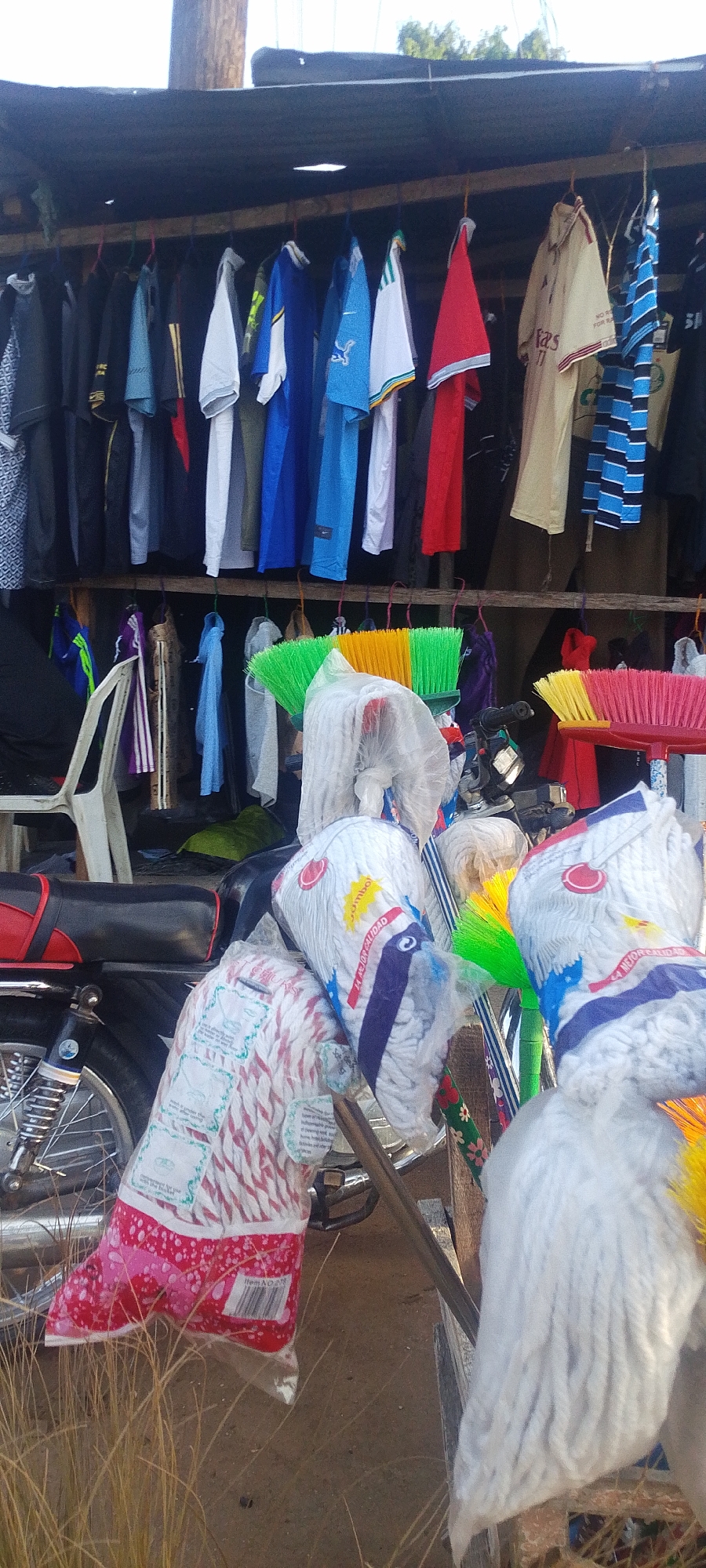 In Lokoja, Nigeria, an outdoor market stall displays a variety of items for sale. Hanging from a rack are numerous articles of clothing, including shirts and jackets. Below the clothing rack, a collection of household cleaning supplies is stacked, including mops in plastic packaging and brooms with brightly colored bristles. A motorcycle with a red and black seat is partially visible behind the cleaning supplies. A white plastic chair is also present in the scene. Dry grass is visible in the foreground. The scene is captured in daylight.