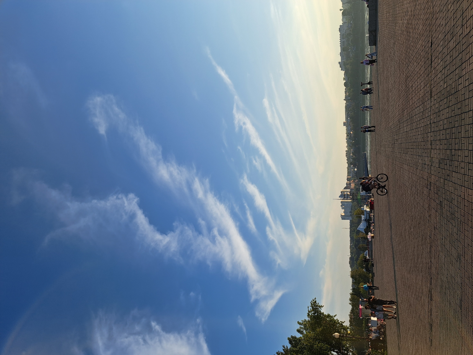 This image depicts a wide, paved promenade along a river in Irkutsk, Russia. The promenade is made of brick pavers and is lined with trees on the left side. Several individuals are visible in the scene. In the foreground on the right, a person is riding a bicycle. Further left, a group of people are gathered, including a family with children. In the mid-ground, a few individuals are walking or standing. In the background, across the river, a cityscape with buildings and trees is visible on a hillside. The sky is a bright blue with scattered wispy clouds, indicating a clear day. The lighting suggests it is late afternoon or early evening, with the sun casting long shadows across the promenade.