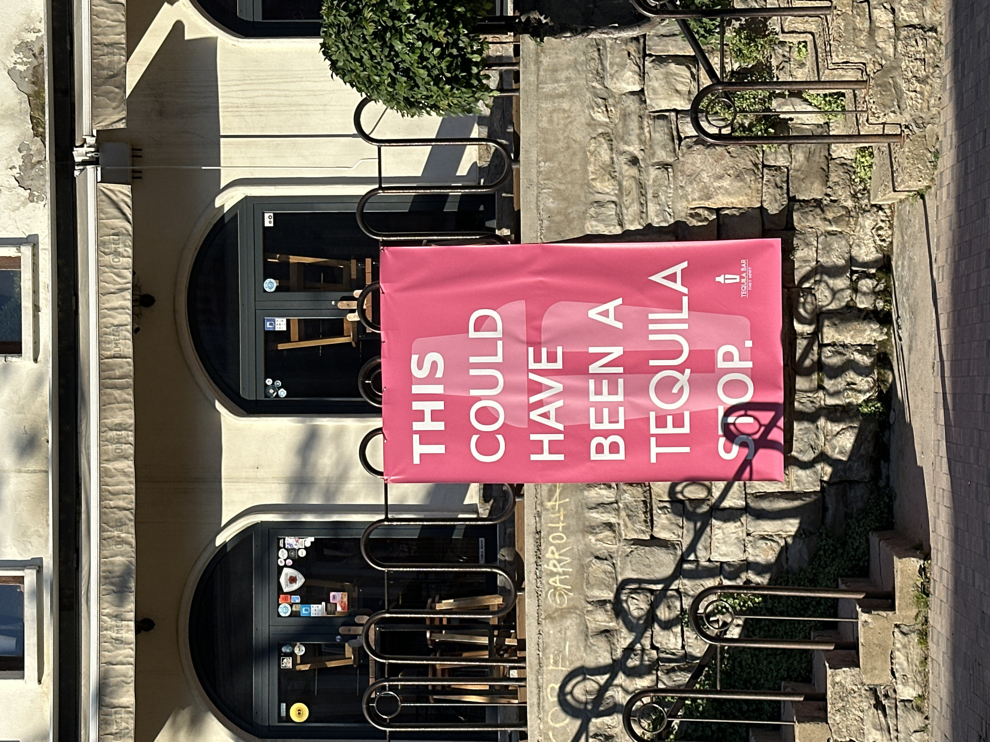 The photograph shows the exterior of a building with two arched windows on a light-colored facade. A large pink banner hangs in front of the building, reading "THIS COULD HAVE BEEN A TEQUILA STOP." A logo on the banner indicates "TEQUILA BAR / TREAT ONLY." Stone steps and a metal railing run alongside the building, with graffiti faintly visible on the stone. The scene appears to be in Veliko Tarnovo, Bulgaria, based on other location clues (not present here). Chairs are visible inside the building.