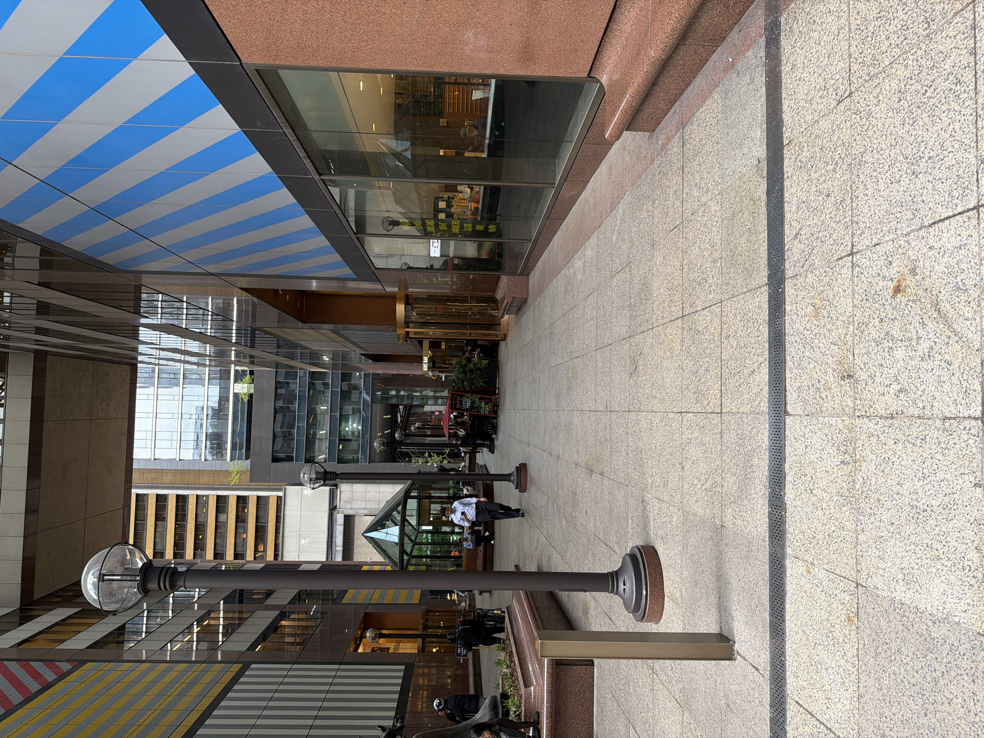 The image captures a pedestrian pathway between modern buildings in an urban setting, identified by visible text as "770 7TH AVE," indicating a location in New York City. The path is paved with light-colored, speckled granite tiles, featuring a long, dark drain grate running down its middle.
Overhead, a blue and white striped canopy extends, providing cover. The building on the left side of the path is predominantly glass, reflecting the surrounding architecture. The building on the right has a facade of reddish-brown granite, with ground-level storefronts enclosed in large glass windows. Within these windows, reflections show street activity, and interior views suggest a business, possibly a cafe or restaurant, with some unreadable text and the word "SOUP" discernible on a menu or sign.
Several individuals are visible along the pathway. A man in a white shirt and dark pants stands in the mid-ground. Others are seated at outdoor tables on the right, suggesting a dining or relaxation area, while more people are seen walking further down the passage. Modern street lamps with spherical light fixtures are placed at intervals. A sign in the distance includes the words "WINE BAR." The scene is illuminated by natural daylight, indicating a clear day with soft shadows.