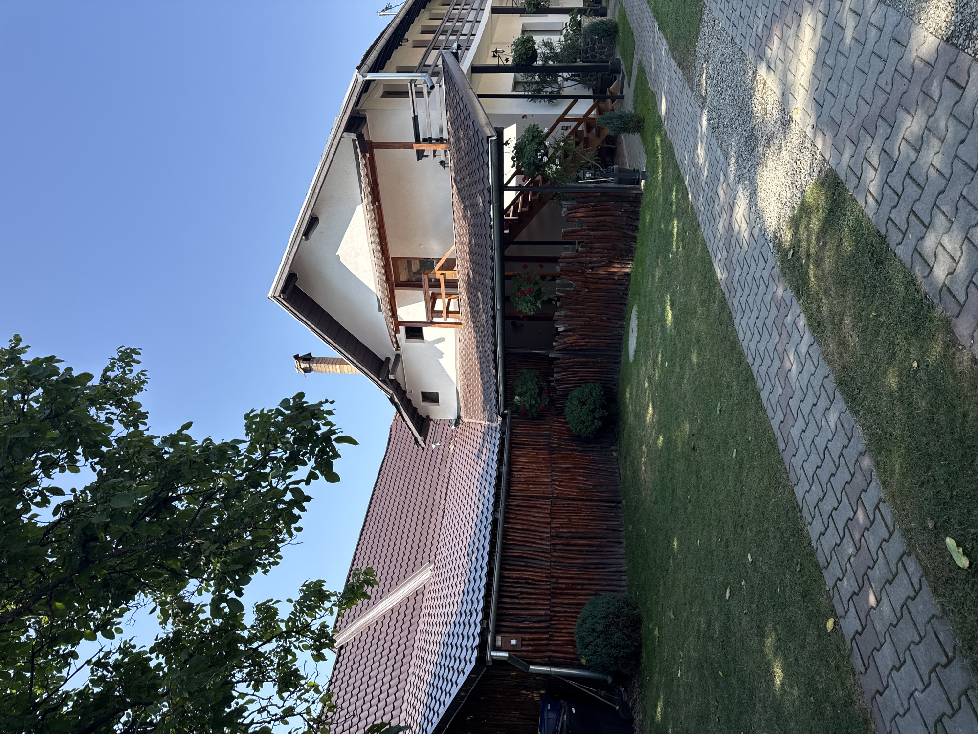 The image depicts a two-story house with a reddish-brown tiled roof, located in Cârțișoara, Romania. The house has white stucco walls and wooden accents on the balconies and eaves. A paved walkway made of interlocking bricks leads to the entrance of the house. The yard is covered with green grass and bordered by a rustic wooden fence. Trees with green leaves are visible in the foreground and partially obscure the view of the house and sky. The sky is clear and blue. The scene appears to be a residential property under a bright, sunny day.