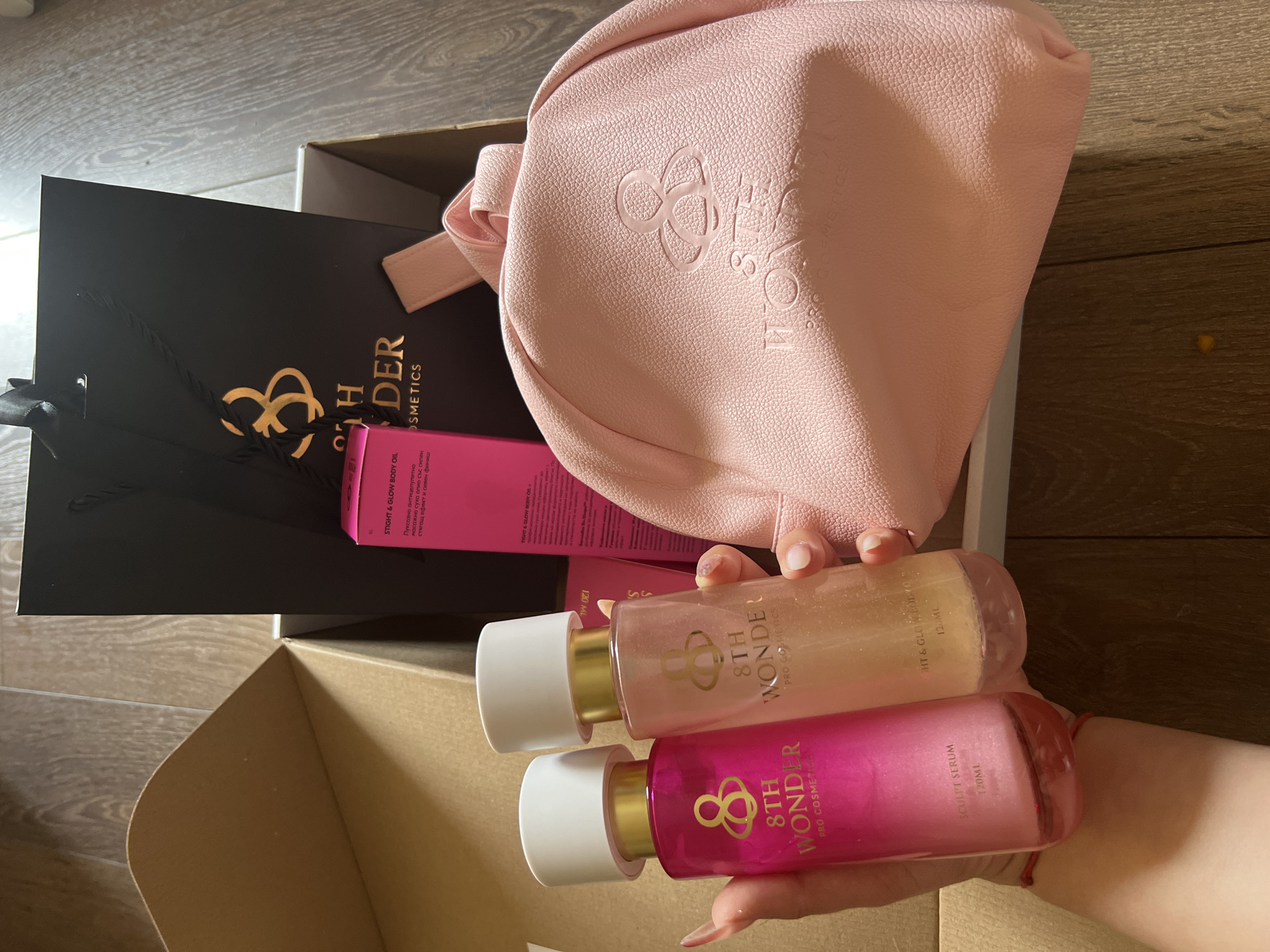 The image depicts a close-up, overhead view of an open cardboard box containing cosmetic products. The box is placed on a wooden floor. Inside the box, a black shopping bag with a gold logo and text is visible. A pink cosmetic pouch with the same gold logo embossed on it is placed on top of some white packaging material. Two bottles of cosmetic serum are held in the foreground by a person's hands. One bottle is bright pink and labeled "8TH WONDER SCULPT SERUM 120ML". The other bottle is clear with a shimmering pink liquid and is labeled "8TH WONDER TIGHT & GLOW BODY OIL 120ML". The person's fingernails are long and painted. A small pink drawstring bag is partially visible. The overall scene suggests a product unboxing or presentation. The location of the image is identified as Sofia, Bulgaria.