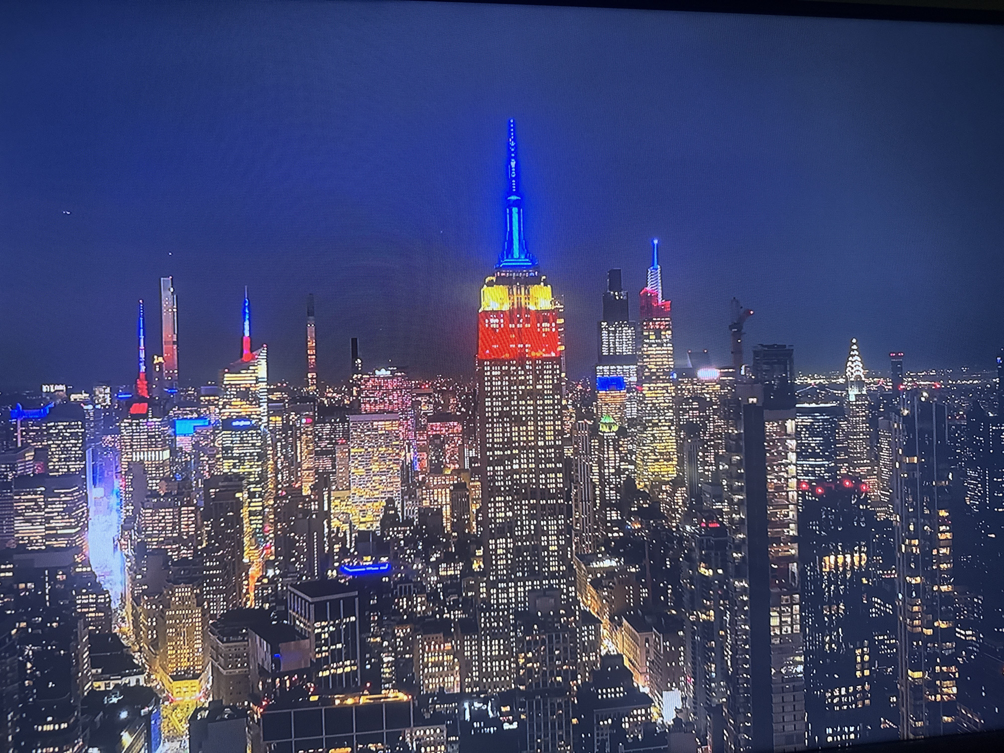 The image depicts the illuminated skyline of New York City at night. The Empire State Building is prominently featured, lit with blue at the top, yellow in the middle, and red at the bottom. Various other skyscrapers are visible, with a mixture of lit windows and colored lights. There are also a few construction cranes visible.
The city is not Varna, Bulgaria.