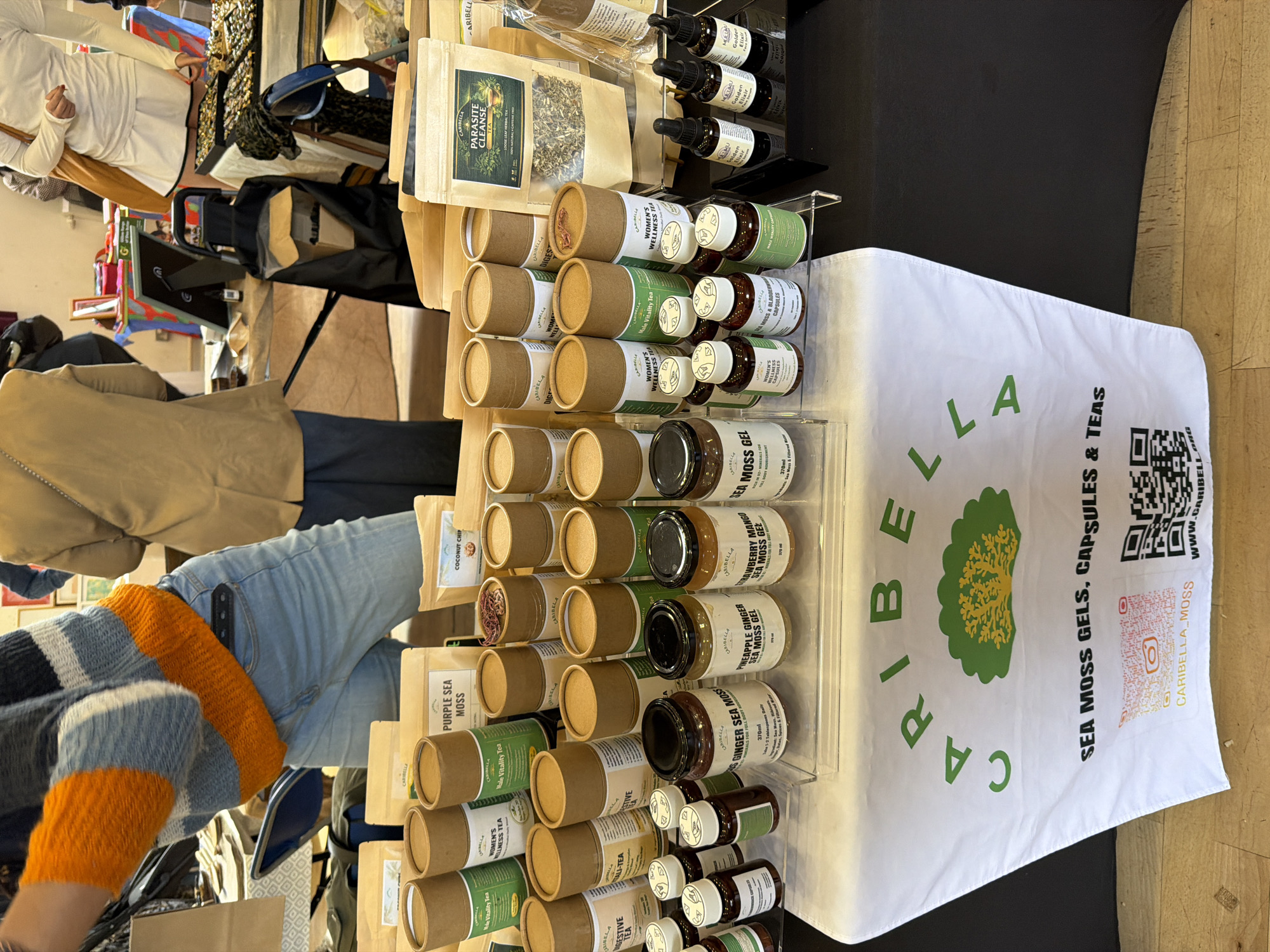 This image captures a commercial presentation, likely at a market or fair, in London, United Kingdom. A table draped with a white cloth displaying the green "CARIBELLA" logo along with "SEA MOSS GELS, CAPSULES & TEAS" is the focus.
The table is densely arranged with rows of health products, including cylindrical cardboard containers and jars of what appears to be sea moss gel, and small bottles with droppers. Products are displayed on clear acrylic stands.
An individual wearing jeans and a blue and orange sweater is partially visible in the lower left corner, presumably interacting with the display. The background is cluttered with other vendor displays and shoppers.