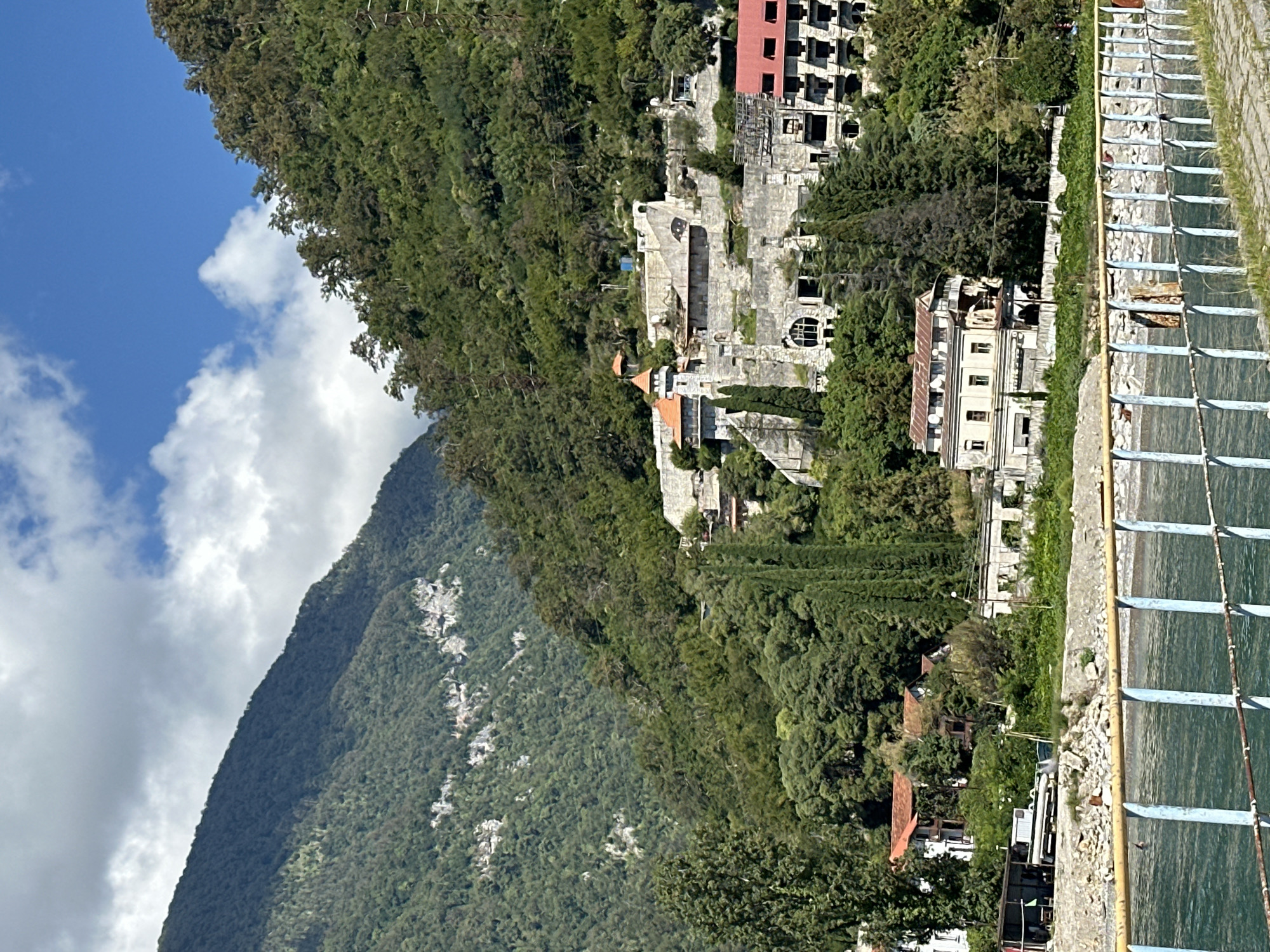A wide shot captures a scenic view of Abaata, Abkhazia, featuring a hillside dotted with buildings overlooking a body of water. Lush green trees and dense foliage cover the steep slopes of the mountains that dominate the background. Several buildings, ranging from older, weathered structures to seemingly abandoned or unfinished ones, are nestled into the hillside.
On the right side of the frame, a large, multi-story building with a red roof appears to be under construction or renovation, with visible scaffolding and unfinished sections. Further up the hill, a complex of stone buildings with a tower and arched windows suggests a historical or unique architectural style. Several smaller houses with terracotta roofs are visible lower on the slope, some appearing occupied.
In the foreground, a blue and yellow striped railing runs horizontally across the image, suggesting a walkway or viewing area along the water's edge. The water itself is a deep teal color, with ripples indicating a gentle current. The shoreline is rocky, with some debris visible. The sky is a bright blue with scattered white clouds, casting shadows on the mountainside. The overall impression is one of a picturesque, somewhat rugged landscape with a mix of natural beauty and human habitation, some of which appears in disrepair.