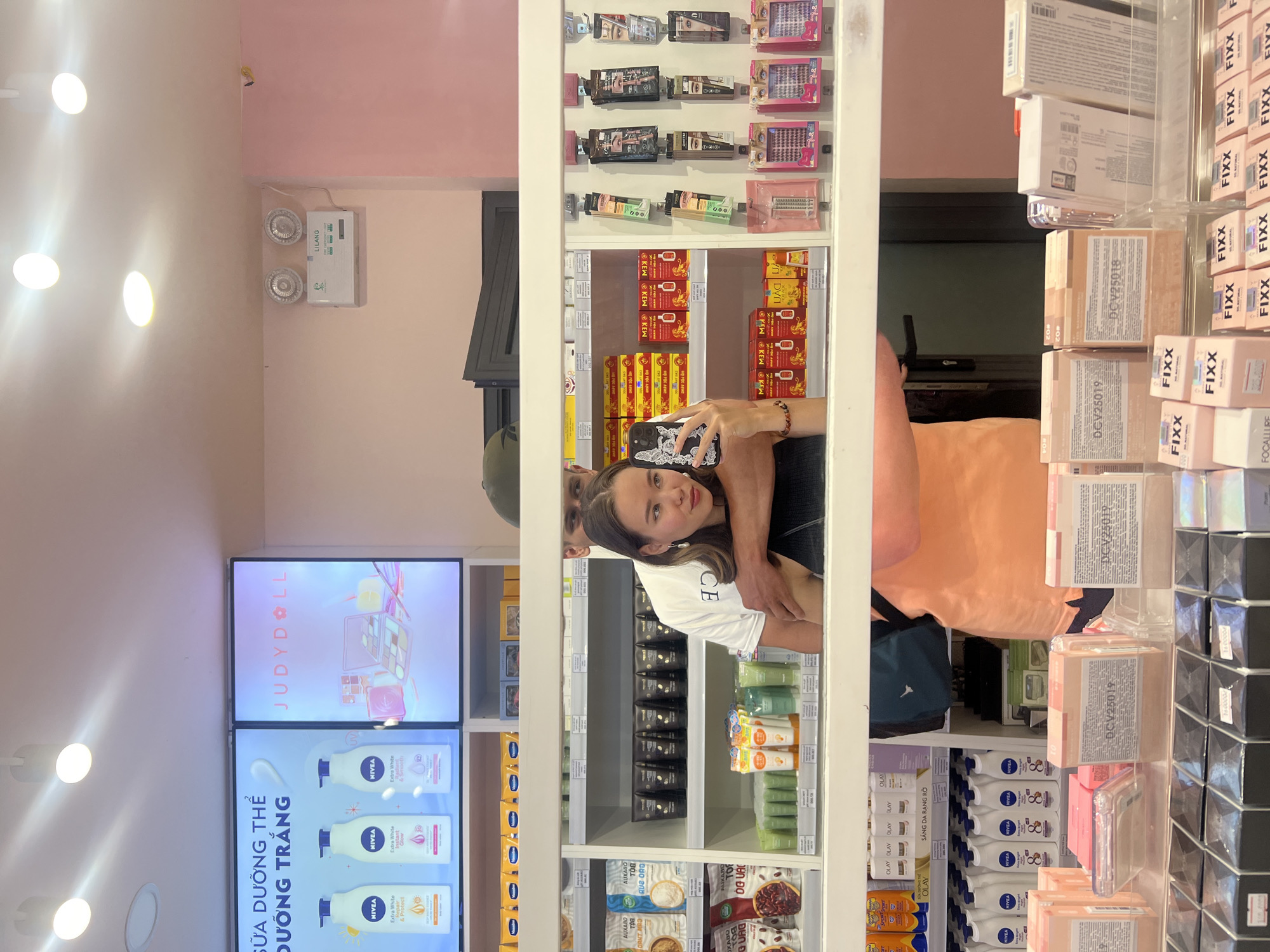 Here is a detailed description of the image:
**Scene Overview:**
The image appears to be taken inside a store, possibly a cosmetics or personal care shop, in Nha Trang, Vietnam. The camera lens has captured the reflections in mirrors or glass doors of shelving units.
**People:**
There are reflections of two individuals in the glass:
1. A young woman holding a mobile phone, presumably taking the picture. She has light skin and is wearing a white t-shirt.
2. An individual standing next to the woman, wearing an orange t-shirt.
**Subjects & Objects:**
* Shelves filled with various cosmetic and personal care products. These products include:
* False eyelashes
* Creams/lotions (some labeled "KEW")
* Skincare products (NIVEA lotions are prominent)
* Makeup products by brands like "FIXX" and "FOCALLUIRE".
* "Auxabo"
* "Olay" products
* Advertisements on a digital display screen for "JUDYDOLL" and "NIVEA" products.
* A white box with the text "LILANG" on it.
* Lighting fixtures.
**Actions & Interactions:**
* The woman is taking a photograph of herself, possibly with her companion reflected in the glass.
* The people are standing near the shelves of products.
**Environment:**
* The store has a pink wall in the background.
* The store is well-lit with fluorescent lights.
* It is a modern, well-organized retail environment.
The location seems to be Nha Trang, Vietnam, based on the text "SỮA DƯỠNG THỂ DƯỠNG TRẮNG" on the digital display.