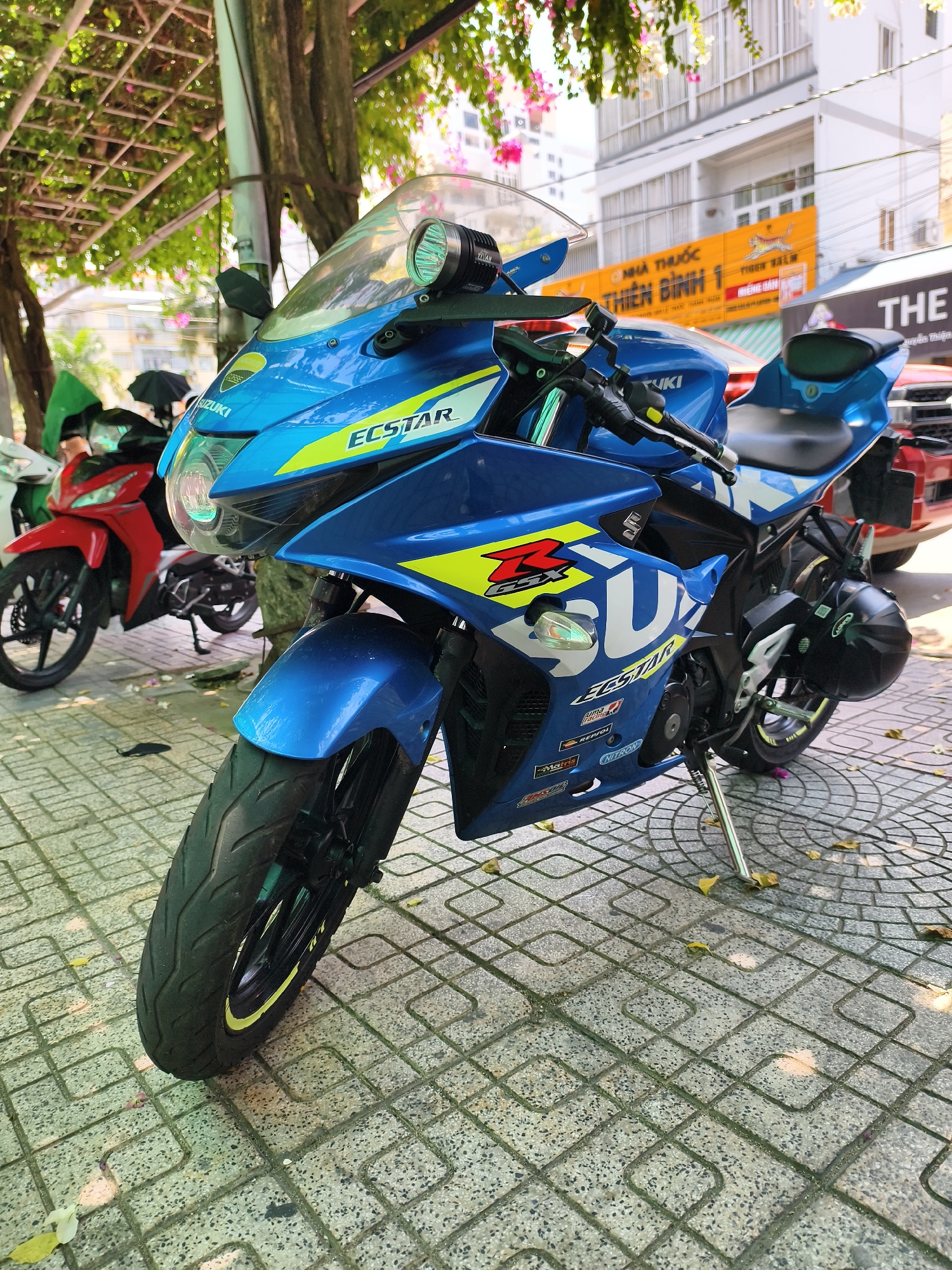 The image captures a blue and yellow Suzuki GSX-R motorcycle parked on a patterned tile sidewalk in Nha Trang Ward, Vietnam. The motorcycle, prominently displayed, has the word "ECSTAR" in yellow and the Suzuki logo on its fairings. A headlight is mounted above the front fairing. In the background, a red scooter, also parked on the sidewalk, is partially visible. Behind the motorcycle, there's a red car and a building with an orange sign that reads "NHÀ THUỐC THIÊN BÌNH 1." A sign reading "THE" is also visible. The scene is framed by trees, one of which is to the left of the motorcycle.