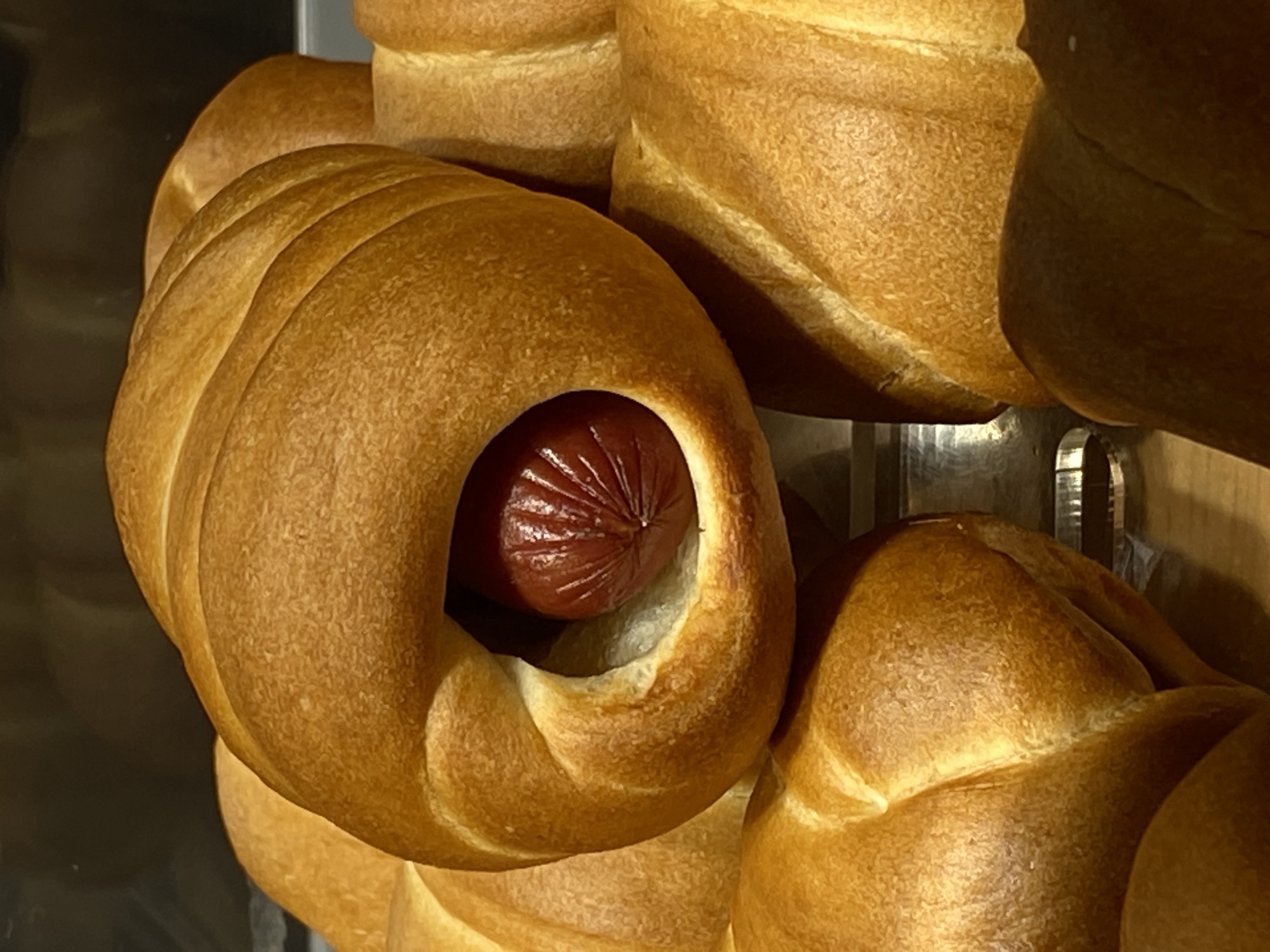 A close-up shot shows a stack of pretzel buns, one of which has a hotdog inserted into the hole. The buns are golden brown, and the hotdog is reddish-brown with visible ridges. The location in Votkinsk, Russia is not discernible from the image.