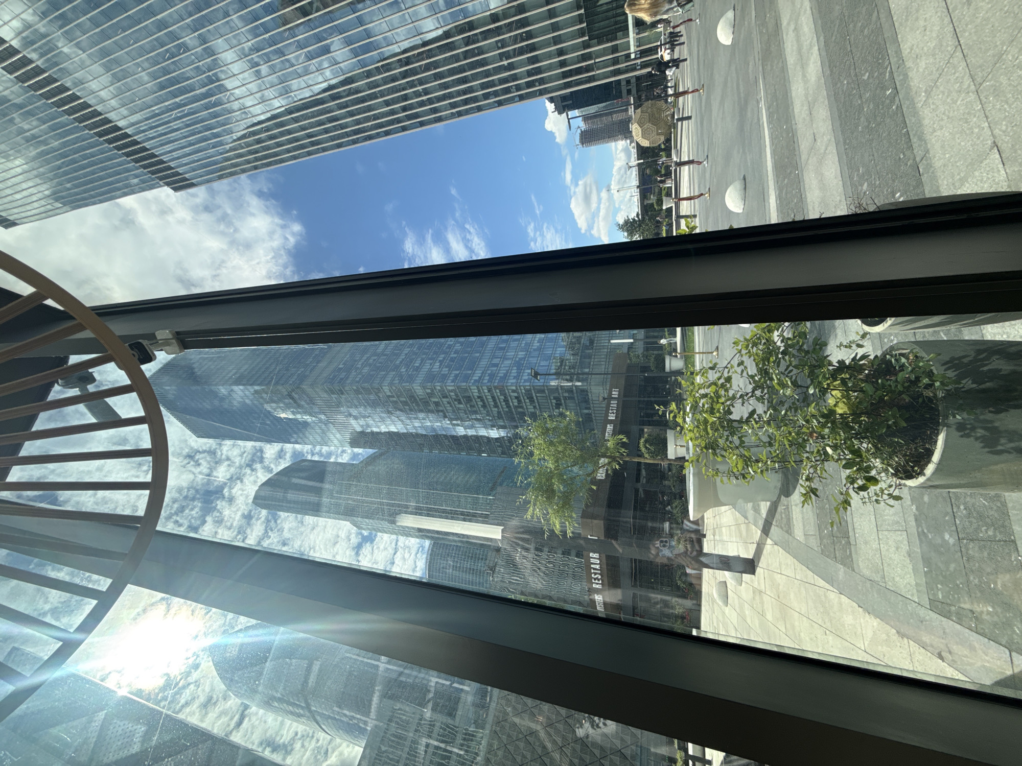 Captured from an indoor vantage point, the image displays an external view of what is located in Khimki, Russia. Skyscrapers dominate the scene, their glass facades reflecting the blue sky dotted with scattered clouds. A clear view of the blue sky and clouds is visible between the buildings. Adjacent to the skyscrapers, a pedestrian walkway is lined with decorative objects, including half-spherical sculptures and trees in pots. The interior perspective is framed by a dark gray window frame. A partial view of a chair with wooden slats is visible. Glare from the sun can be seen on the glass.