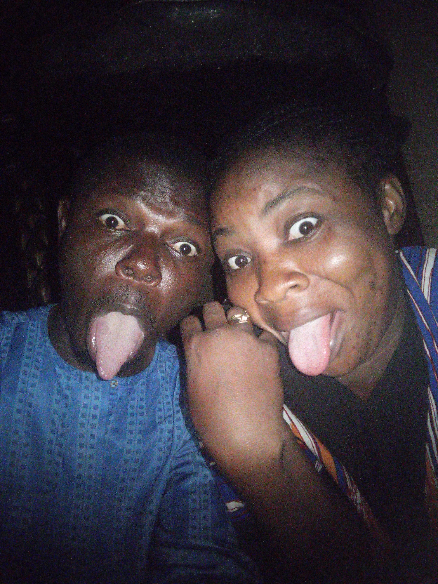 Two individuals, one male and one female, are shown in a close-up, head-and-shoulders shot, both sticking out their tongues. The man on the left is wearing a blue patterned shirt. The woman on the right is wearing a black shirt with a colorful striped garment layered over it and a ring on her left hand. The background is dark and appears to be indoors. There are no discernible landmarks or environmental features to suggest a specific location within Tempure, Nigeria.