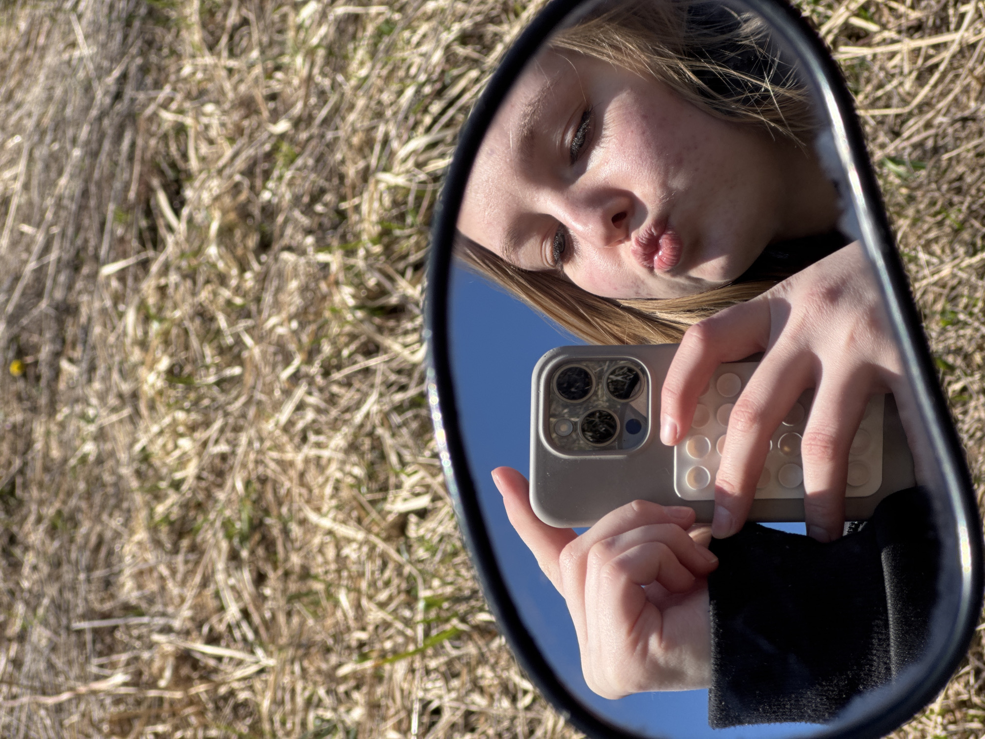 A person is taking a selfie in a mirror. The person is holding a smartphone with a gray case. The person is making a kissing face. The background shows a field of dry grass and a blue sky, reflected in the mirror. The person is wearing a black top. The location is Zhigalki, Russia.