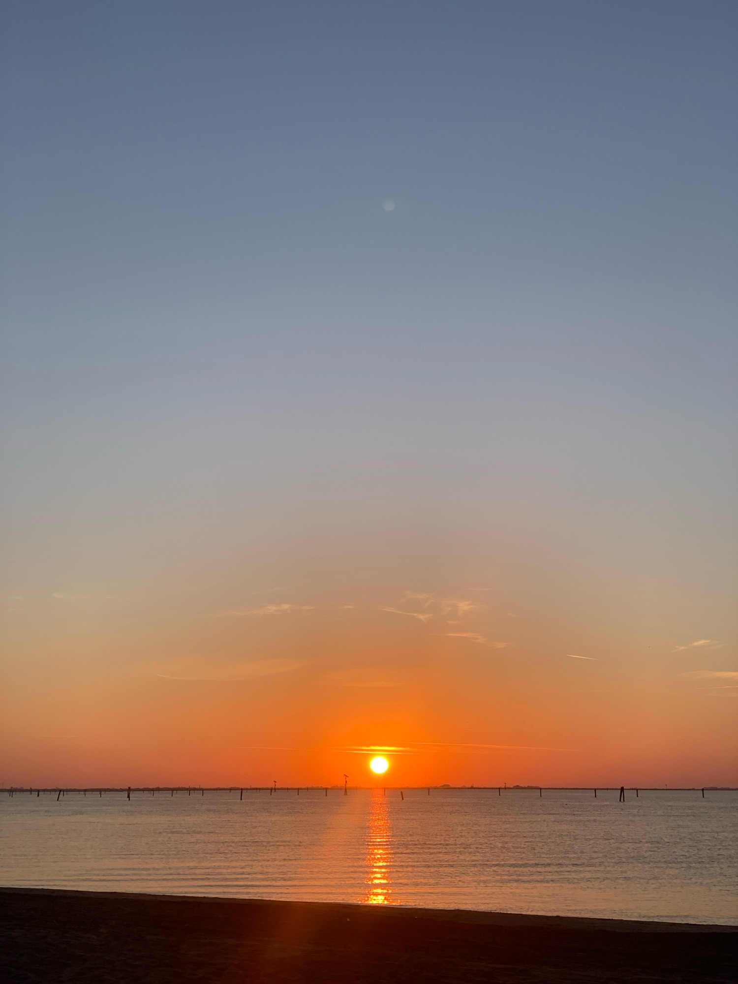 The image depicts a sunset over a calm body of water, likely the Adriatic Sea, near the city of Comacchio, Italy. The sun, a bright orange orb, is low on the horizon, casting a shimmering reflection across the water. Numerous posts, likely marker buoys or part of an ancient fishing system, are visible in a line across the water. The sky transitions from a pale blue at the top to warm oranges and pinks near the horizon, with a few wispy clouds present. The foreground shows a dark, sandy beach. There are no people or animals visible in the image.
