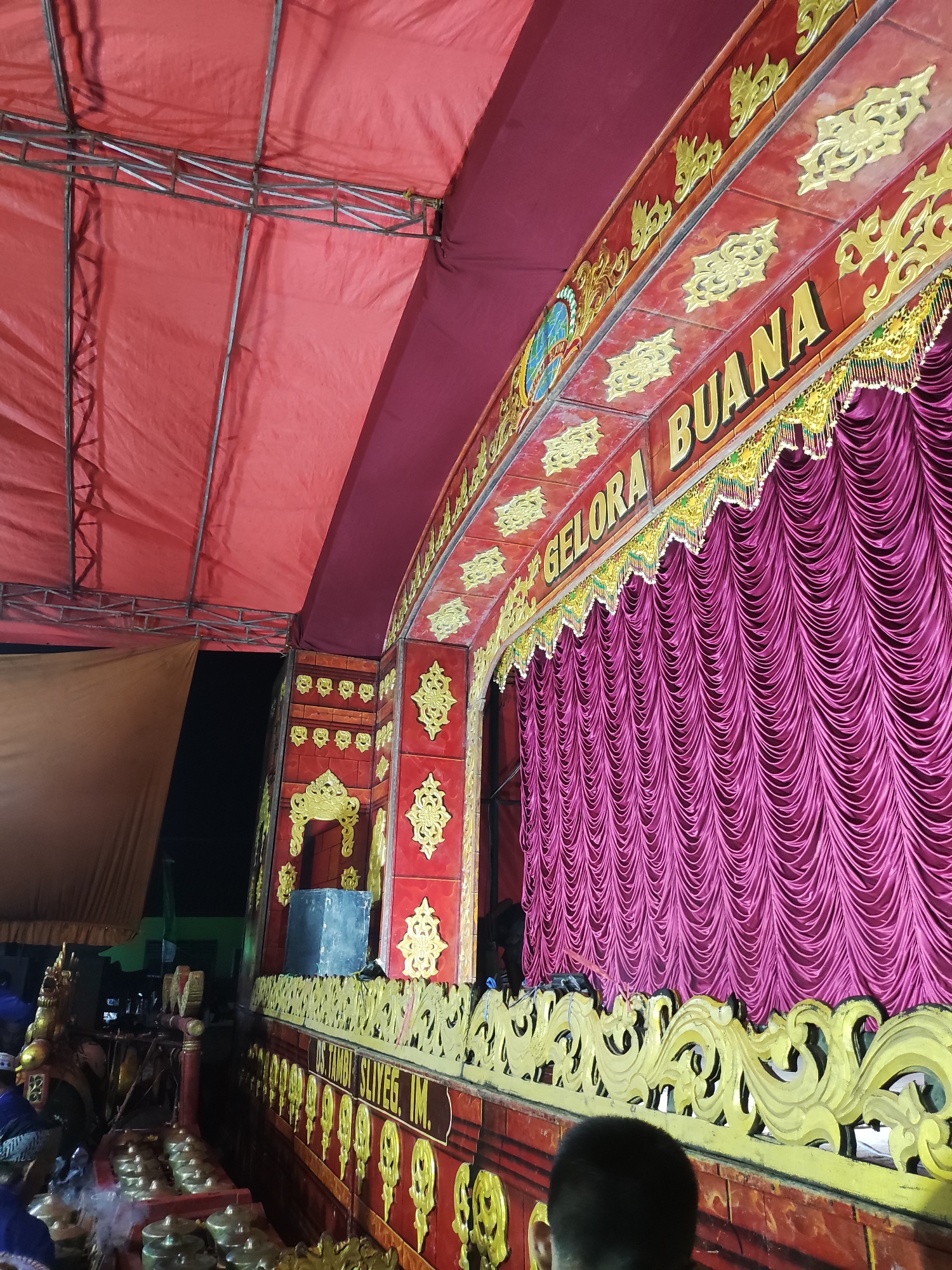 The image depicts a stage set for a performance in Panongan, Indonesia. A large, red tent structure covers the top of the scene, supported by metal scaffolding. The stage facade is elaborately decorated with red panels featuring gold ornamental details. A prominent sign across the top of the stage reads "GELORA BUANA" in large gold letters. Below this, a luxurious purple curtain is drawn, concealing the stage area. In the foreground, to the left, part of a gamelan instrument ensemble is visible, consisting of bronze percussion instruments arranged on red stands. Several individuals, their faces obscured, are present near the instruments. To the right, the back of a person's head is visible in the foreground, looking towards the stage. The overall scene suggests an outdoor cultural or traditional performance is about to commence or has recently concluded.