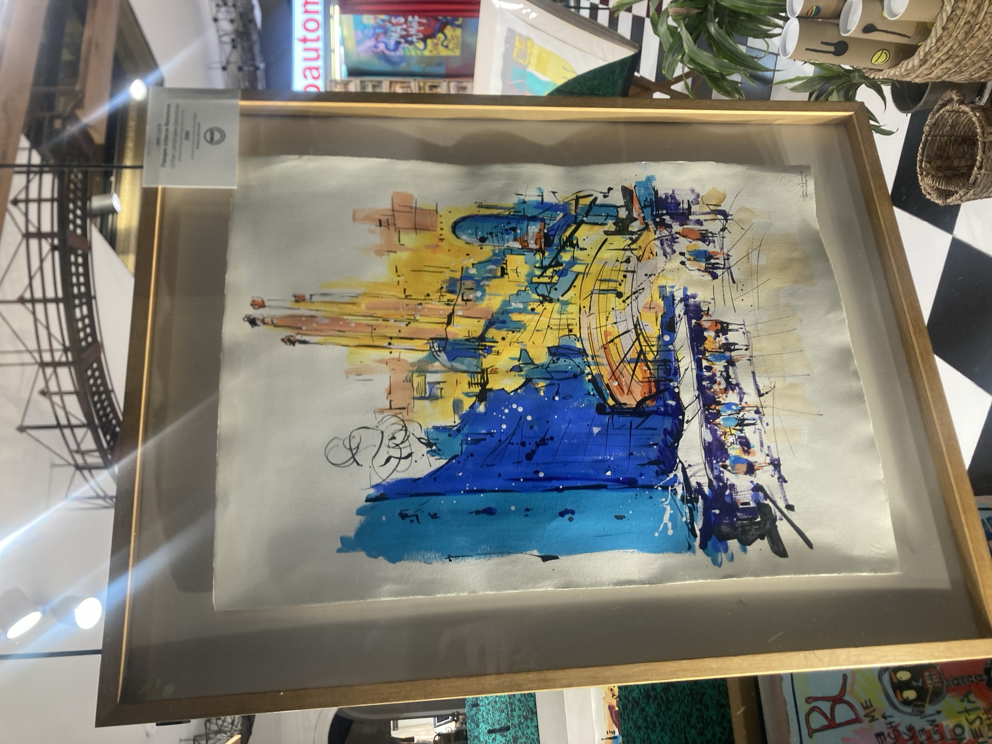 The image shows a framed watercolor painting displayed in an art gallery or exhibition. The painting depicts a vibrant, abstract cityscape with tall, spire-like buildings rendered in bold strokes of yellow, orange, and blue. Figures of people walking along a street are visible in the foreground, also rendered in an impressionistic style. The painting appears to be signed by the artist in the bottom right corner.
Behind the painting, a sign with text is visible, and part of the text reads "Paisaje Urbano Barcelona" which translates to "Urban Landscape Barcelona." This strongly suggests that the painting depicts a scene from the city of Barcelona, Spain. The architecture in the painting, with its distinctive spires and colorful hues, is reminiscent of Antoni Gaudí's work, particularly the Sagrada Familia.
The exhibition space features lighting fixtures from the ceiling and a metal structure that resembles a bridge or elevated walkway in the background. The floor is a black and white checkered pattern. To the right of the painting, there are potted plants and woven baskets, suggesting a retail or display area. Other artwork, including a framed piece with the letters "BL" and a character resembling a robot, is visible to the left. A rack of cylindrical objects, possibly prints or posters, is also present in the background. The overall impression is a dynamic and colorful artistic representation of an urban environment in Barcelona.