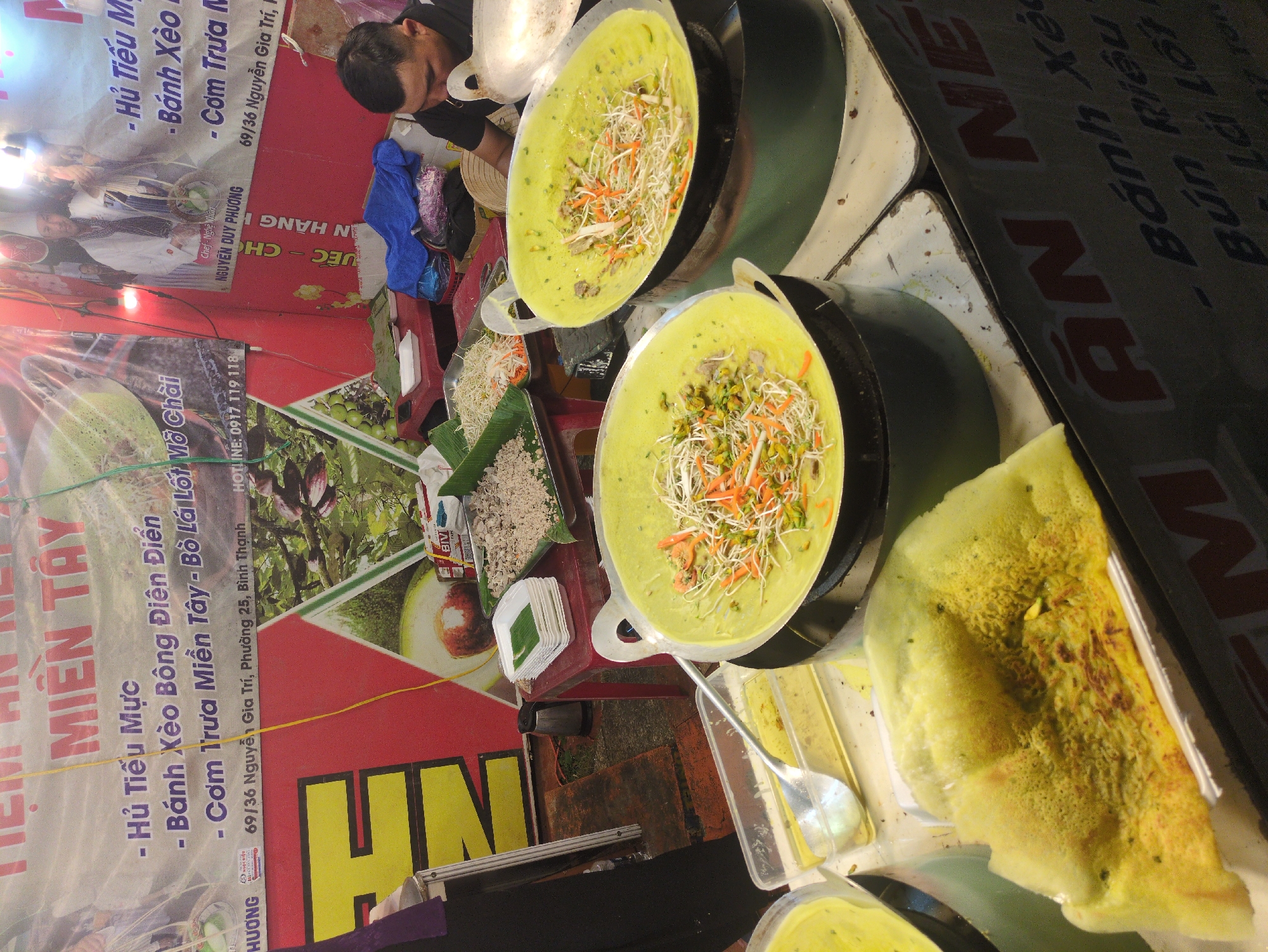 A street food vendor in Tuy Hòa, Vietnam prepares Banh Xeo, a savory crepe. A vendor in a black shirt is working over a cooking station with two pans of pale yellow batter topped with vegetables like shredded carrots and sprouts. To the side is a tray of cooked ingredients, perhaps the filling for the Banh Xeo. A folded Banh Xeo rests on the counter, ready to serve. Banners advertise the street food stall "Tiệm Ăn NE". A menu lists dishes like "Hủ Tiếu Mực" (Squid Noodle Soup) and "Cơm Trưa Miền Tây" (Southern Vietnamese Lunch). The address listed is "69/36 Nguyễn Gia Trí, Phường 25, Bình Thạnh." The area is likely crowded and bustling.