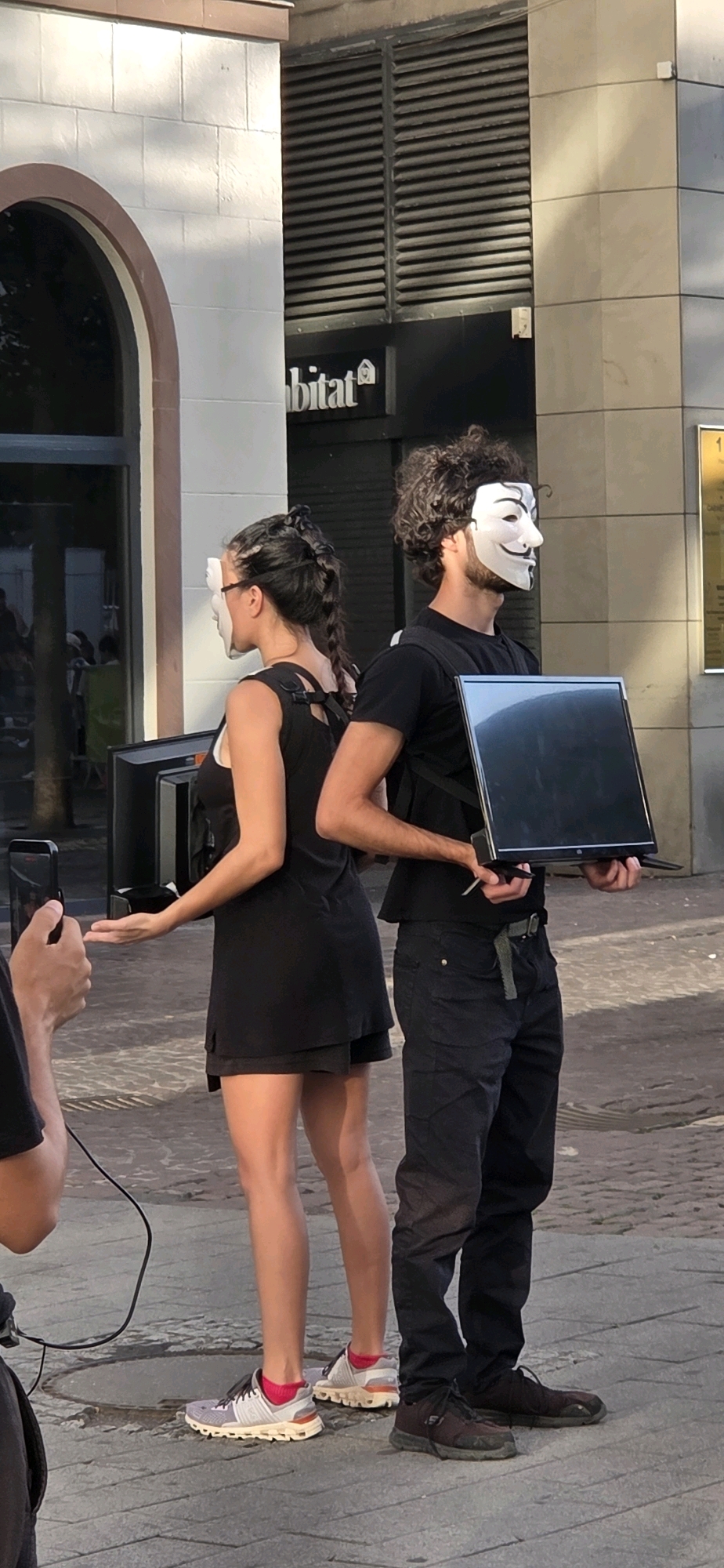 Two individuals, a woman and a man, stand back-to-back in an urban setting, both wearing white Guy Fawkes masks. The woman, with her hair in a braid, wears a black sleeveless dress and pink-accented athletic shoes. She holds her left hand out, palm up. The man, with curly hair, wears a black t-shirt, dark pants, and dark athletic shoes. He holds a black laptop in his hands. Behind them, a large building features a sign that reads "habitat". The scene appears to be a public square with cobblestone pavement.
