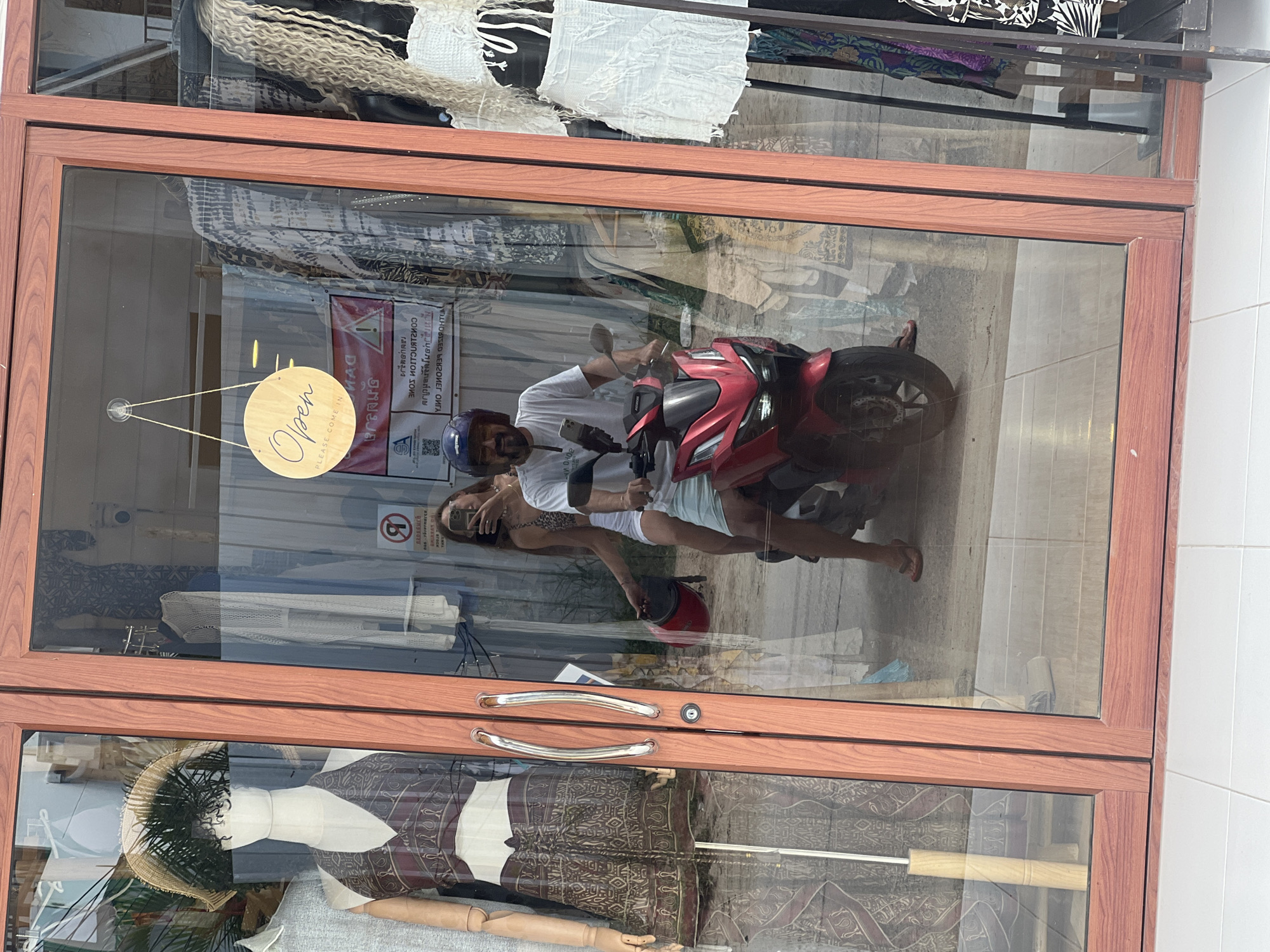 The image captures a reflection in the glass storefront of a boutique in Koh Phangan, Thailand. A man wearing a white t-shirt and blue helmet is riding a red scooter, with a woman seated behind him holding a red helmet. The woman is wearing a leopard-print top and white shorts, and is taking a selfie. The storefront displays mannequins wearing fashionable clothing, and racks of garments are visible inside the shop. A circular "Open, Please Come In" sign hangs from the ceiling. The overall scene suggests a bustling commercial area on the island.