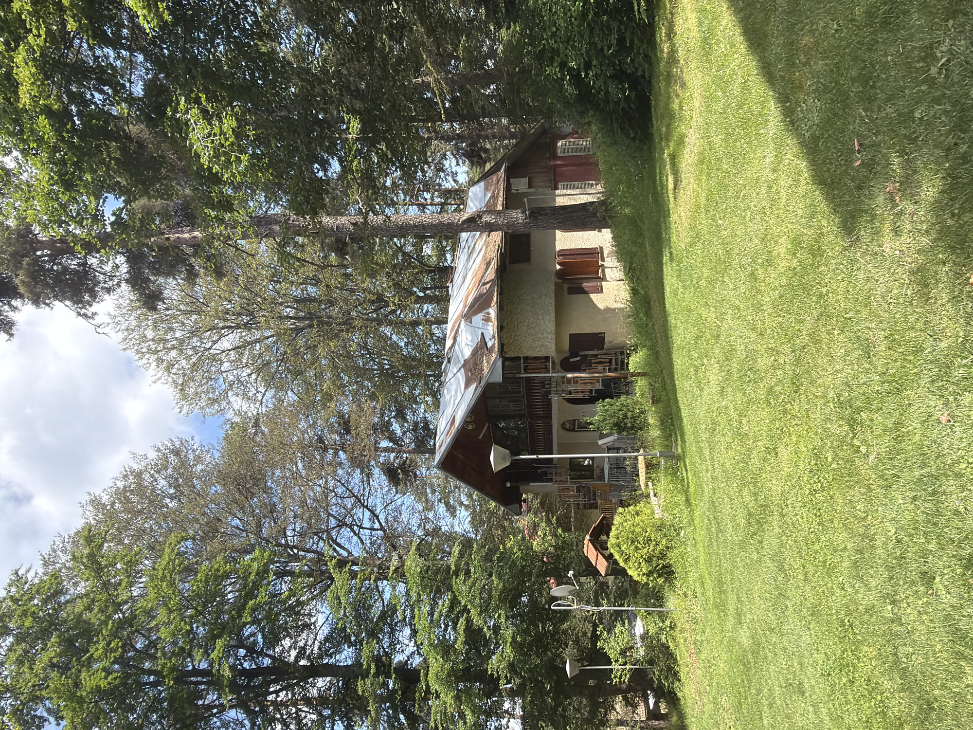 A two-story house with a rusty metal roof is situated in a wooded area. The house is a light tan color with dark brown shutters and a balcony on the second floor. There are trees of varying heights surrounding the house, with a large grassy lawn in the foreground. The sky is partly cloudy.