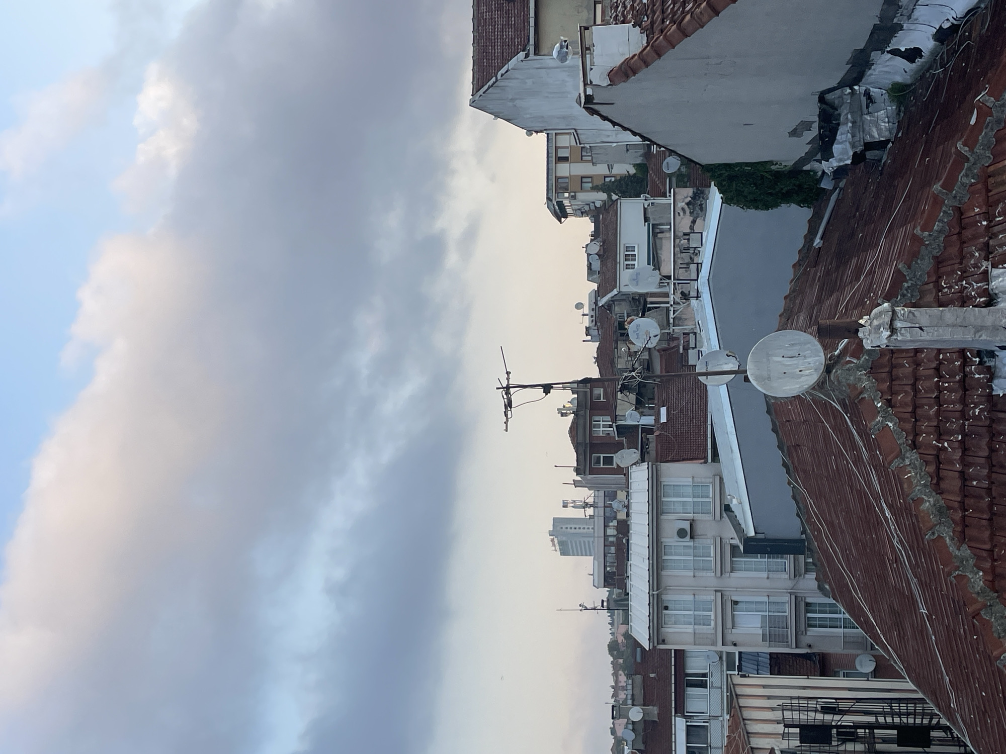 This image depicts a view of rooftops and buildings in the city of Kadıköy, Turkey. The foreground is dominated by a reddish-brown tiled roof with visible aging and repair work, including sections of concrete and plaster. Several satellite dishes are scattered across this roof and those of adjacent buildings, some appearing weathered.
Behind the immediate foreground, various buildings of different architectural styles and heights are visible. These include multi-story residential buildings with balconies and windows, some featuring white facades, while others have darker brick or stone exteriors. There is a prominent modern, light-colored tall building in the mid-ground, featuring numerous windows. Utility poles and antennas are also present among the structures.
The sky above is partly cloudy, with soft, diffused light suggesting it might be early morning or late afternoon. The overall impression is an urban residential area with a mix of older and newer constructions, characterized by a high density of buildings and visible infrastructure for communication. No people are visible in the frame.
