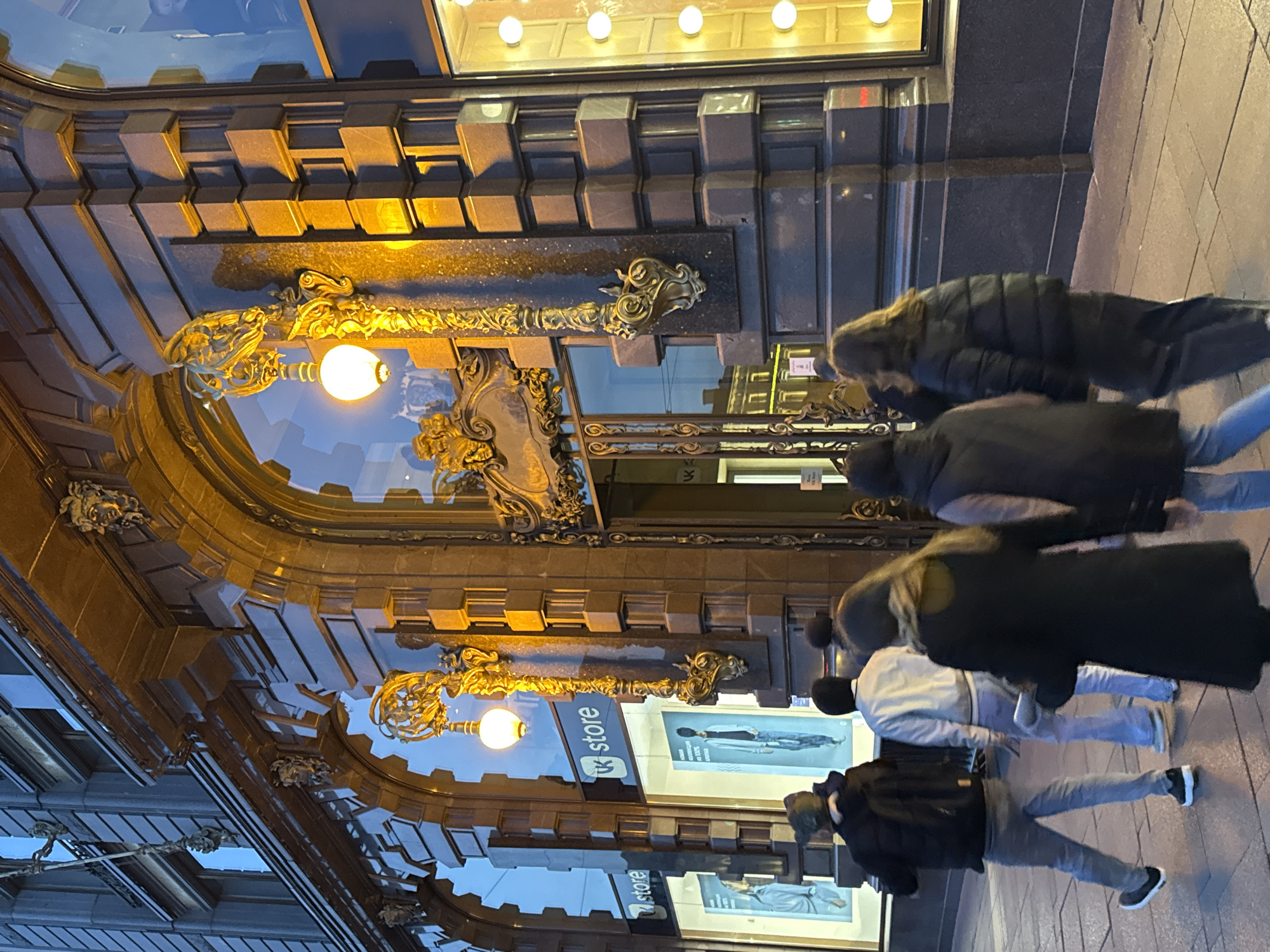 The image shows the Singer House building (also known as Dom Knigi or House of Books) located in Saint Petersburg, Russia. The building features an ornate facade with decorative elements, including elaborate light fixtures and architectural details around the entrance. A storefront with the "VK store" logo and posters of models displaying apparel is visible. Several pedestrians are walking along the sidewalk in front of the building. They are wearing winter clothing, including coats and jackets, and are blurred due to movement. The street and sidewalk appear wet, suggesting it may have recently rained. The time of day seems to be dusk or night, indicated by the lighting from the building and streetlights.
