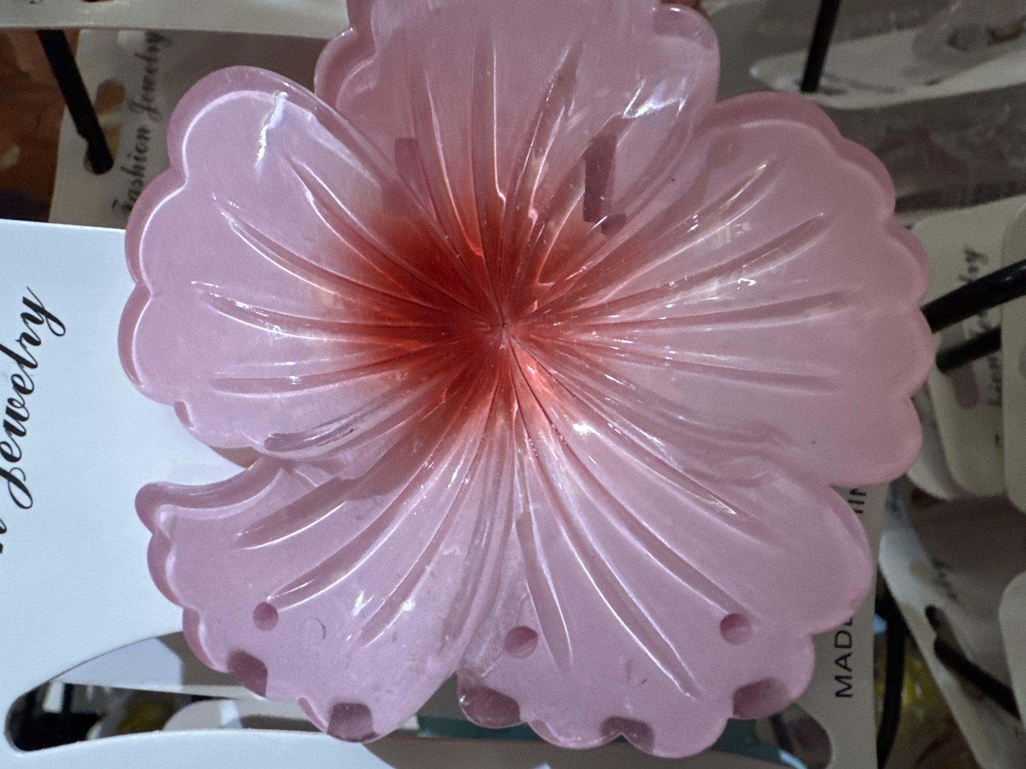 The main subject of the image is a translucent, pink, flower-shaped object, possibly made of plastic or resin. It is intricately molded or carved to resemble a blossom, with distinct radiating lines extending from a central point towards the petals, giving it a textural, grooved appearance. The center of the flower exhibits a deeper reddish-pink hue, which gradually fades to a lighter, softer translucent pink towards the outer edges of the petals. The petals have a scalloped or wavy edge, and several small, circular indentations or holes are visible near the outer tips of some petals.
The flower object appears to be resting on or displayed against white cardstock or packaging. In the background, parts of these white cards are visible, featuring black text. The partial phrase "Fashion Jewelry" can be seen on one card, while another card shows the word "MADE" (likely part of "MADE IN..."). Dark, thin lines, possibly elastic bands or parts of a display rack, are also present in the background. The scene suggests an indoor setting, possibly a retail display for hair accessories or decorative items. No specific activity is taking place; it is a static shot of the object. The lighting appears even, without strong shadows indicating a particular time of day.