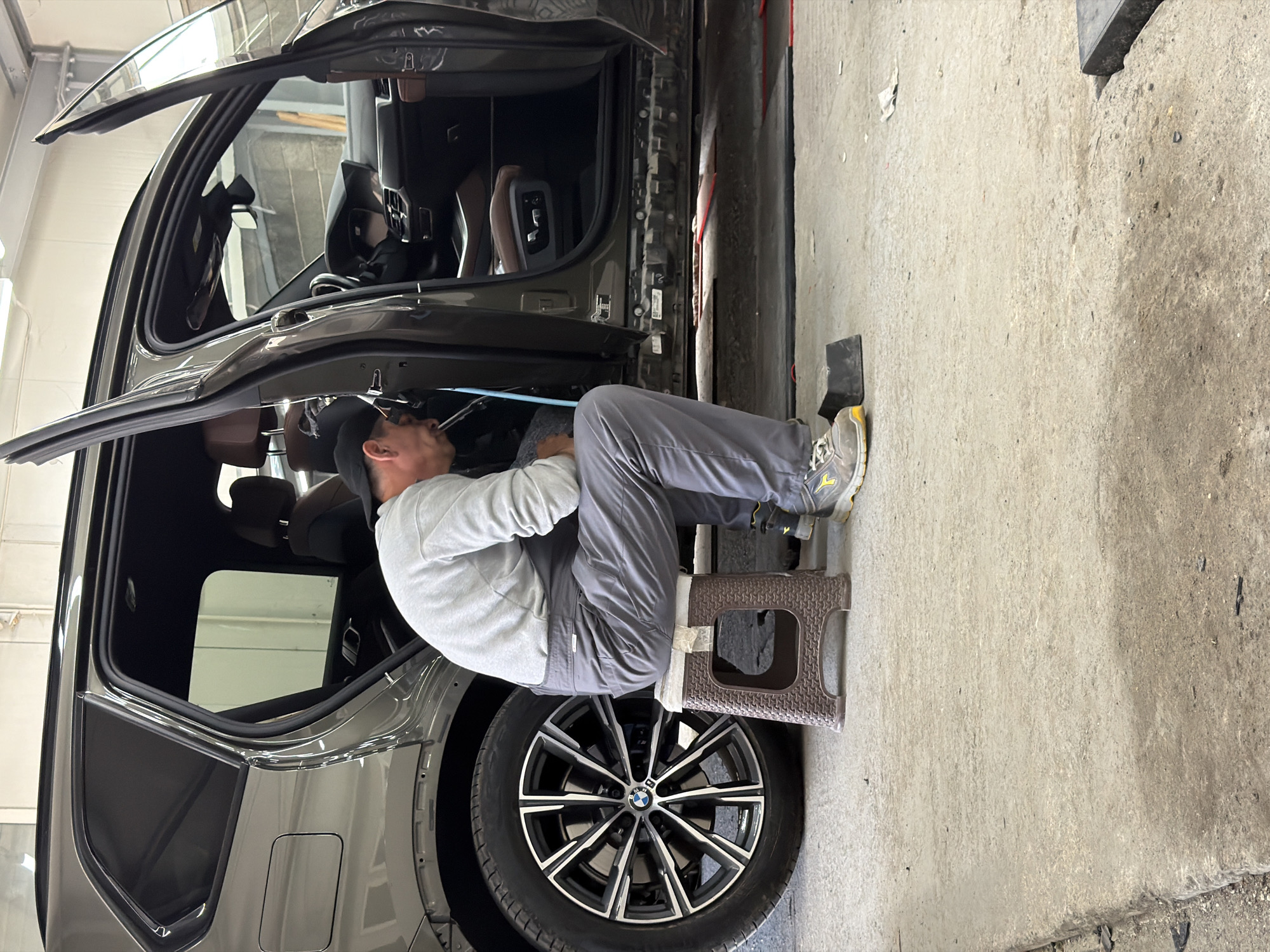 A man is working inside a dark gray BMW SUV, the doors are open, and he is sitting on a small brown plastic stool. He is wearing a gray long-sleeve shirt, gray pants, a black baseball cap, and work boots. He is looking up inside the car. The car is in a garage with a concrete floor and walls. The scene is not explicitly identified as being in Burgas, Bulgaria, but it can be inferred from the available data.