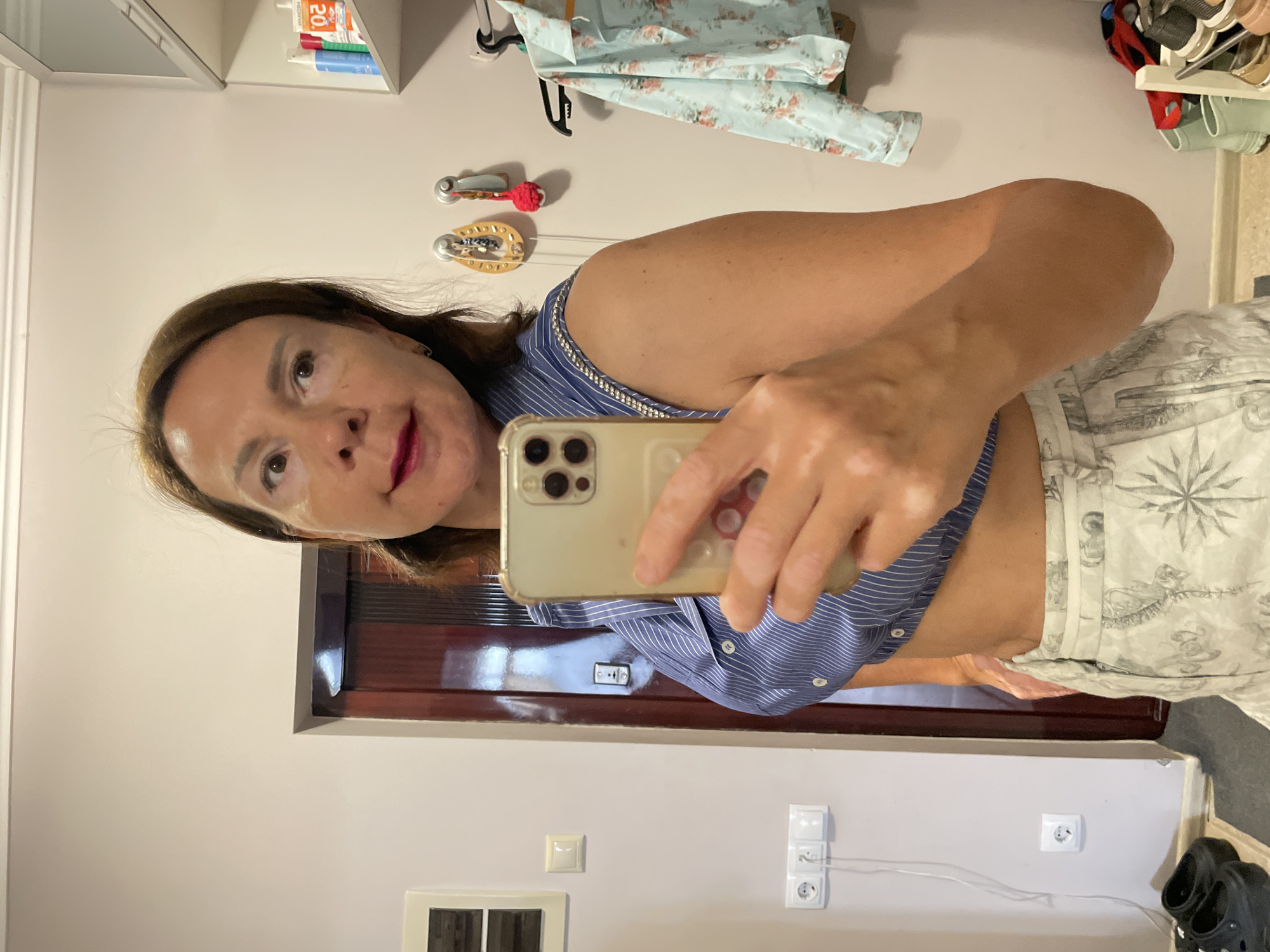 A woman with brown hair and light skin, wearing a blue and white striped crop top and patterned pants, is taking a selfie in what appears to be a domestic interior. Her skin has patches of vitiligo. She is holding a gold iPhone to take the photo, with her right arm extended. In the background, a dark brown door, electrical outlets, and a shelf with toiletries and clothing are visible. The setting is a hallway or closet area within a residence. There are black Croc-style shoes on the floor to the left, and a shoe rack with various footwear on the right.