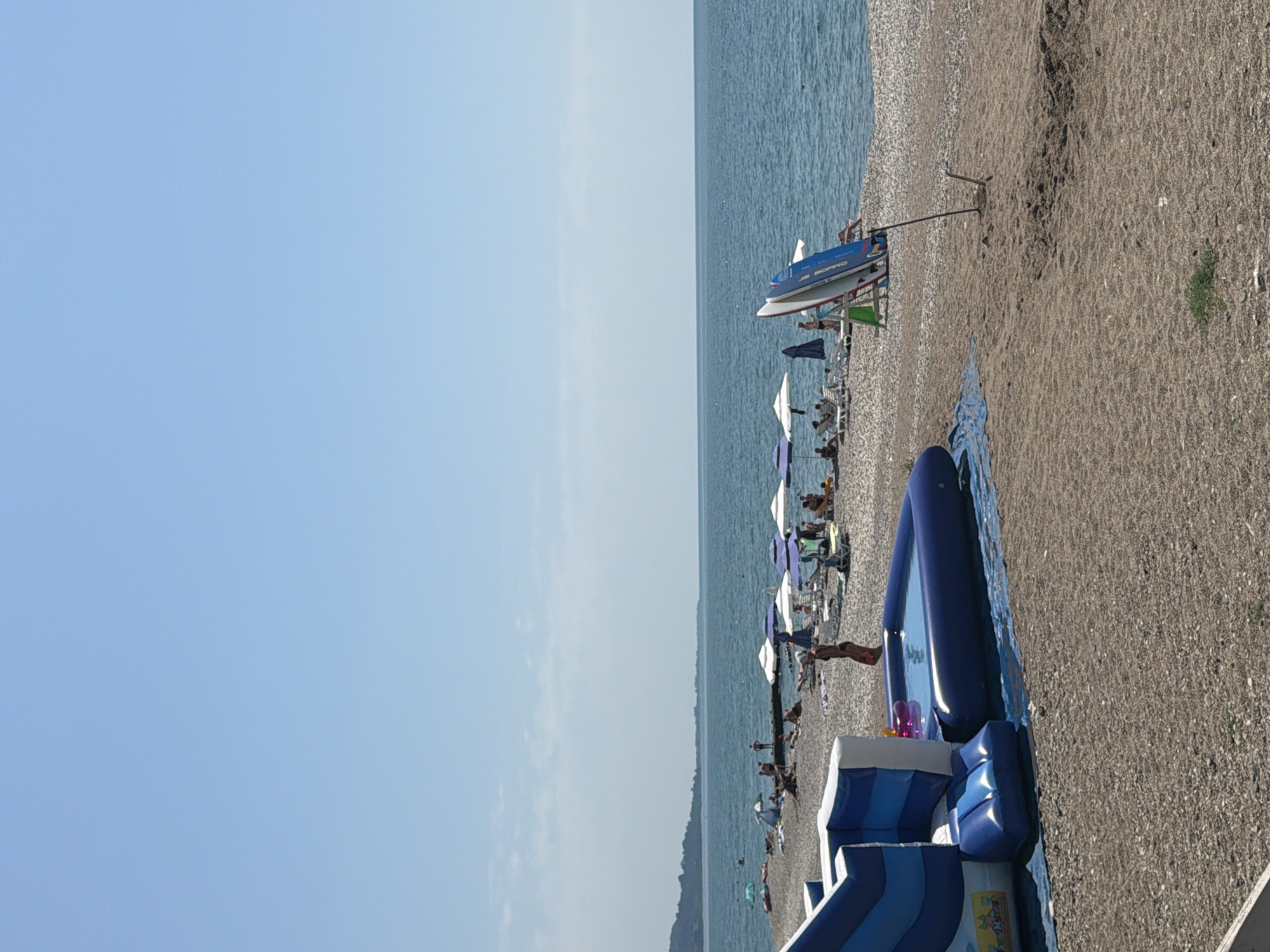 The image depicts a sunny day at a pebble beach in New Athos, Abkhazia. In the foreground, a blue inflatable water slide and a small inflatable pool are set up on the beach. To the right, two paddleboards are propped up against a stand. The middle ground shows a section of the beach populated with sunbathers relaxing under umbrellas, with some individuals in the water. The background features the calm, blue sea stretching to the horizon, with a forested coastline visible in the distance. The sky is clear and blue.